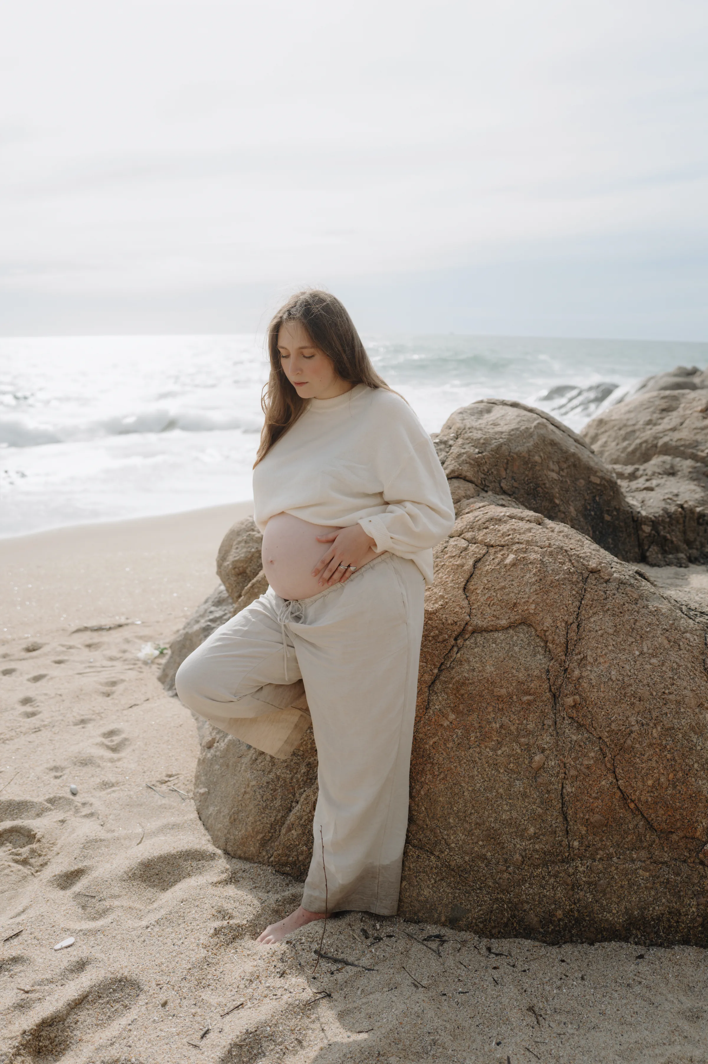Seaside portrait of a person in cream clothing standing by rocks in soft light.