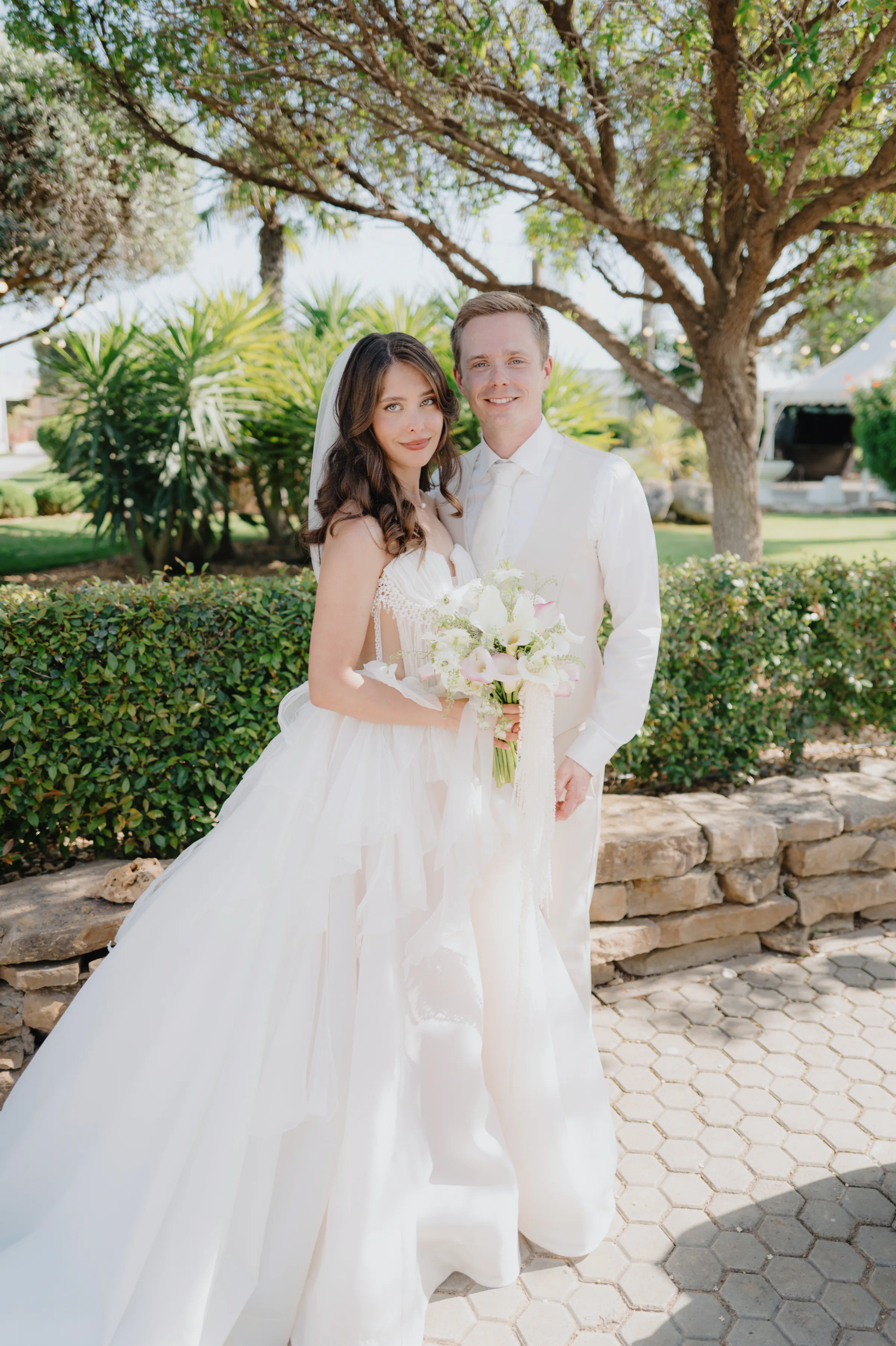 Couple in wedding attire standing in a sunny garden with bouquet and soft light.