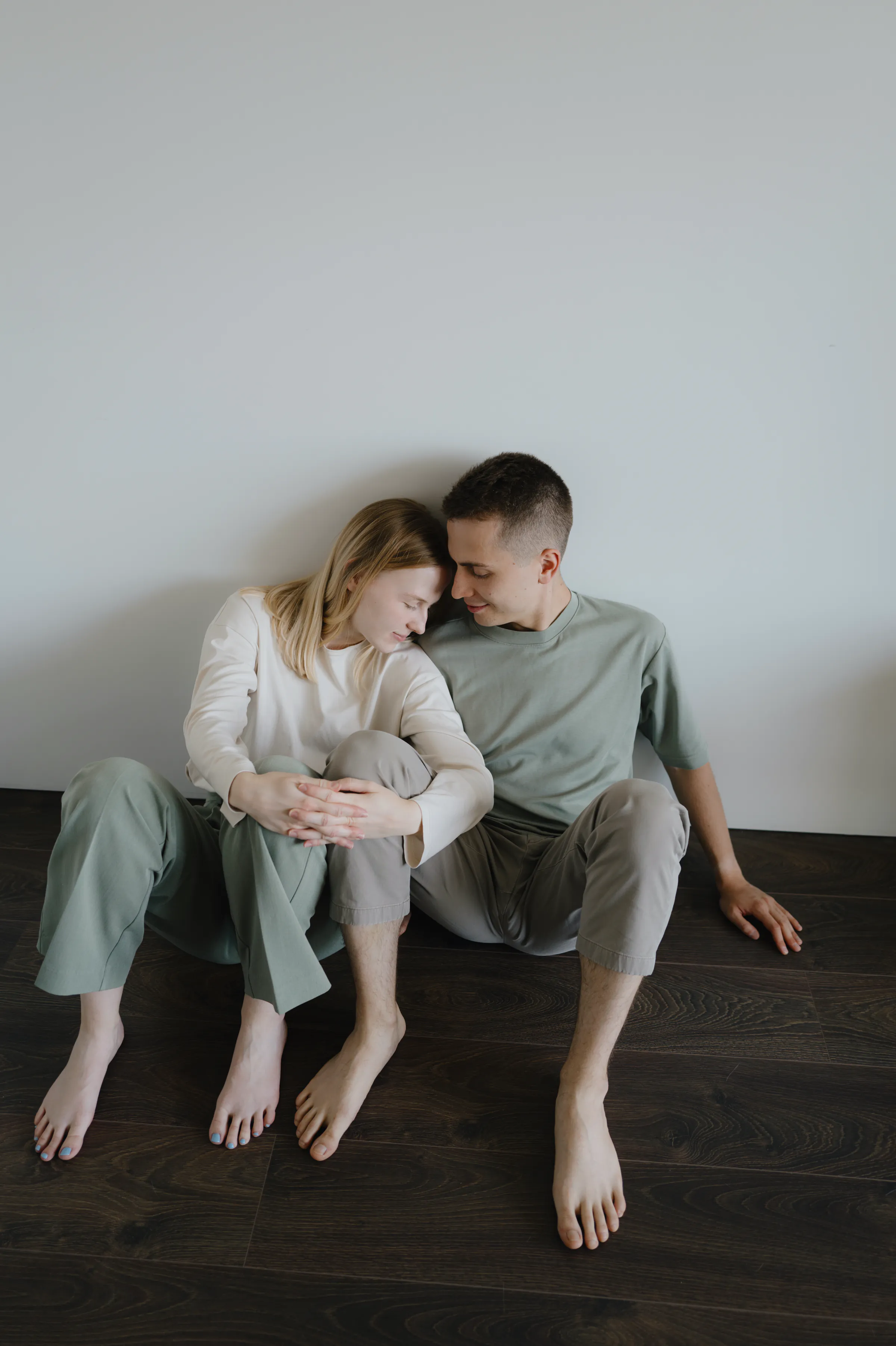 Couple seated barefoot on a dark wood floor against a plain wall in soft indoor light.