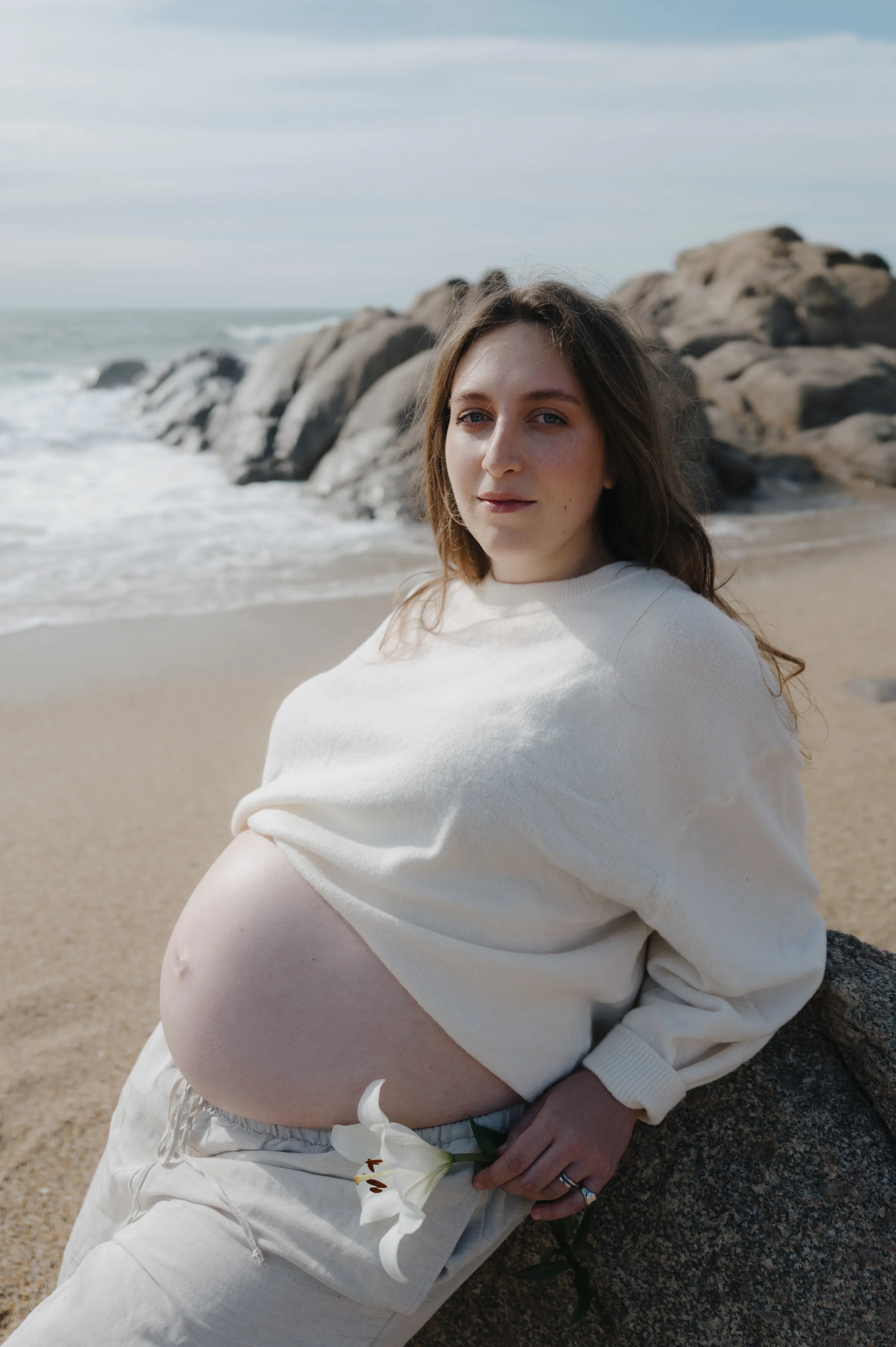 Seaside portrait of a person in white seated by rocks in soft light.