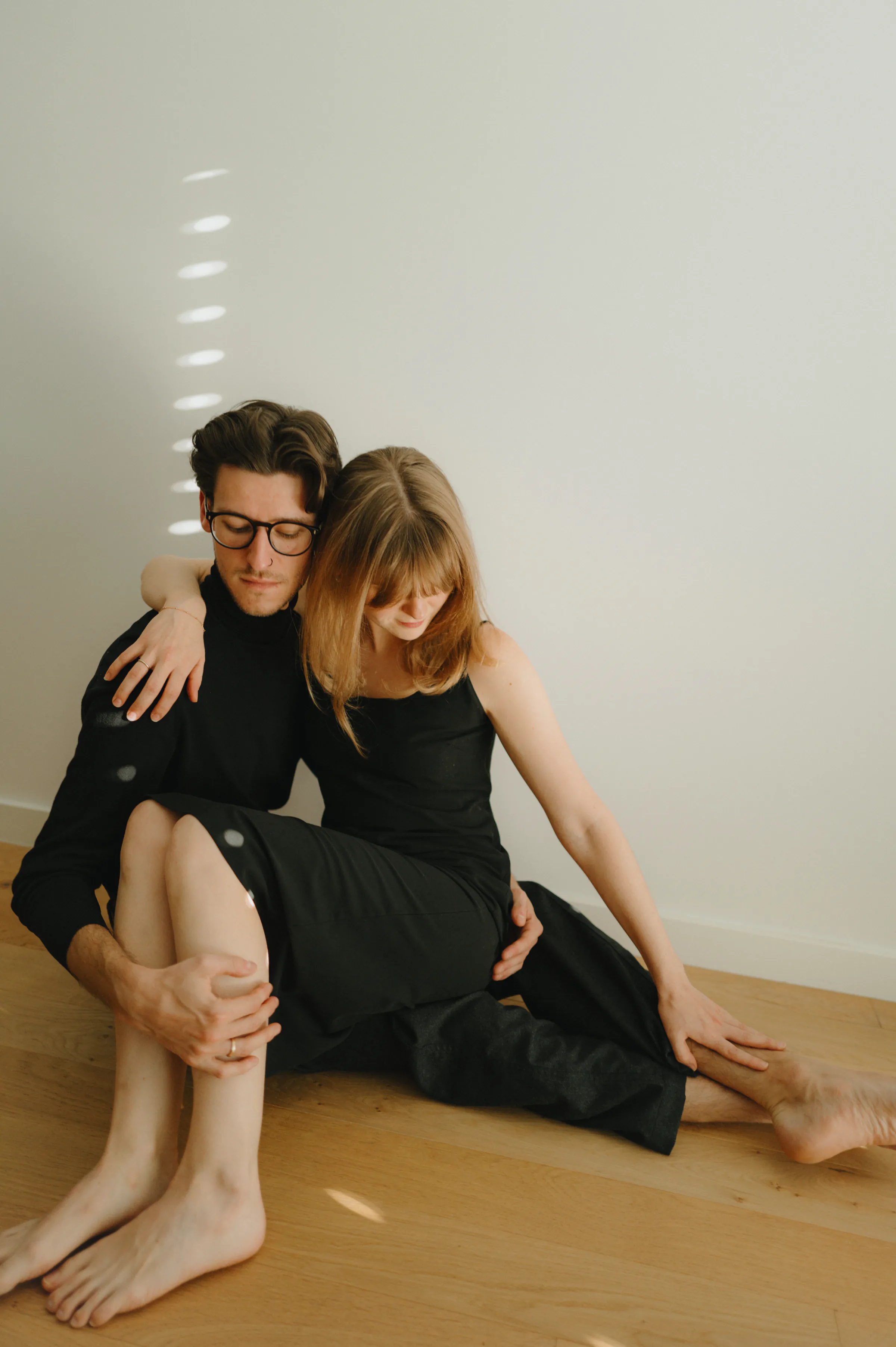 Indoor portrait of a seated couple in black outfits, leaning close against a plain wall in soft light.