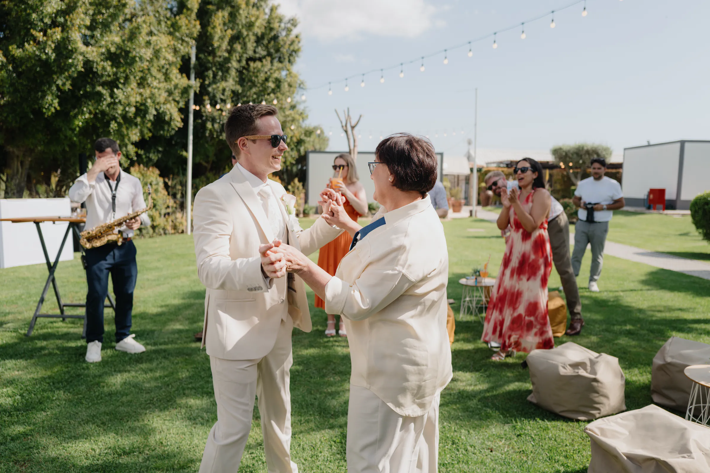 Couple in white suits dancing on a lawn during an outdoor celebration with musicians and guests.