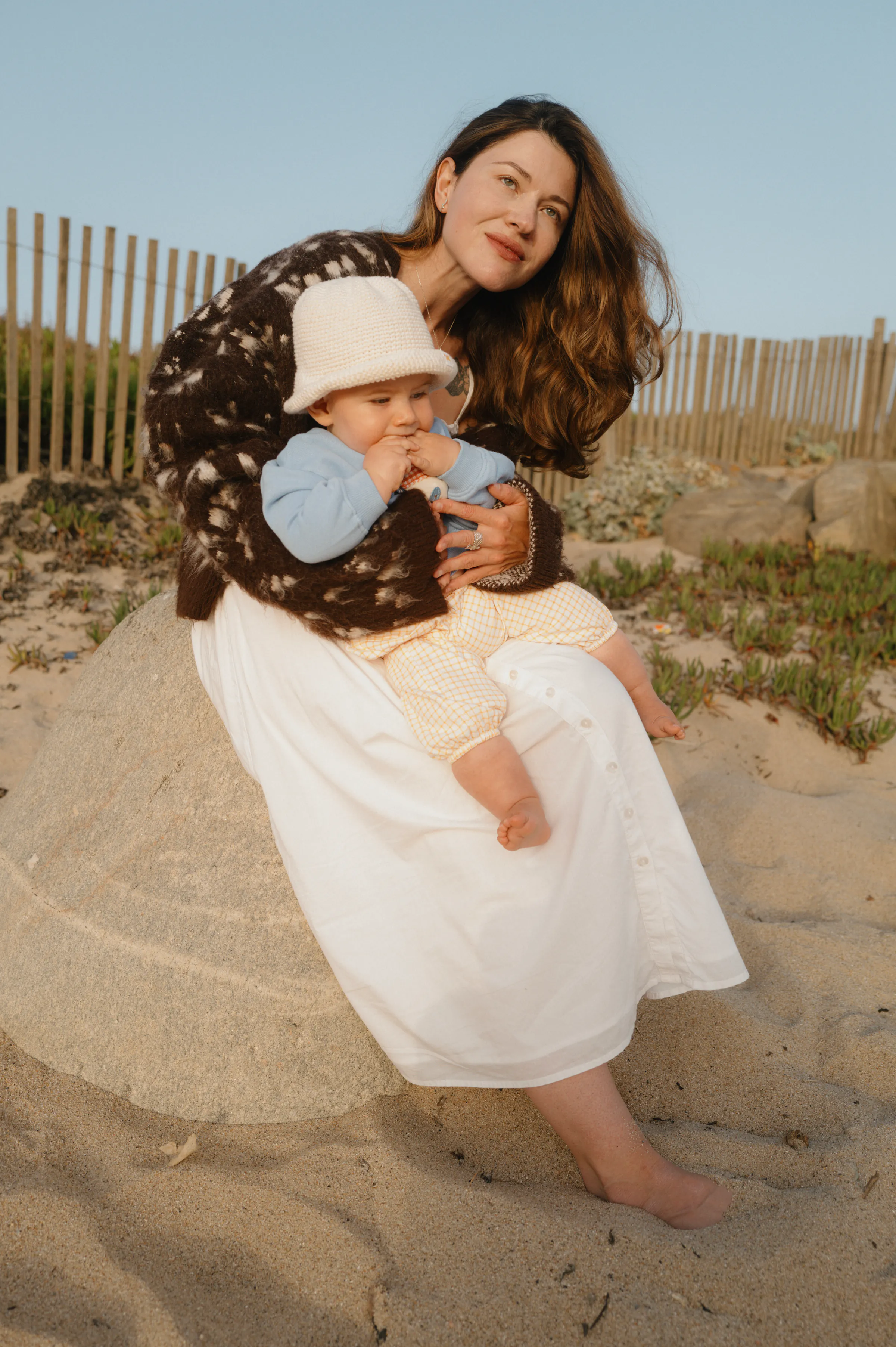 Adult holding a baby during a seaside outdoor session on sandy dunes with beach fencing behind them.