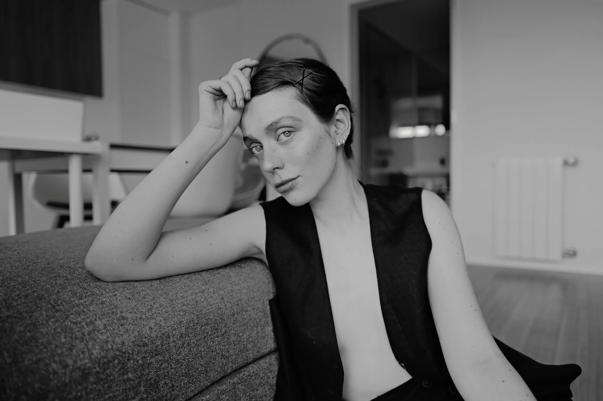 Black-and-white home session portrait of an adult seated beside a sofa in soft indoor light.