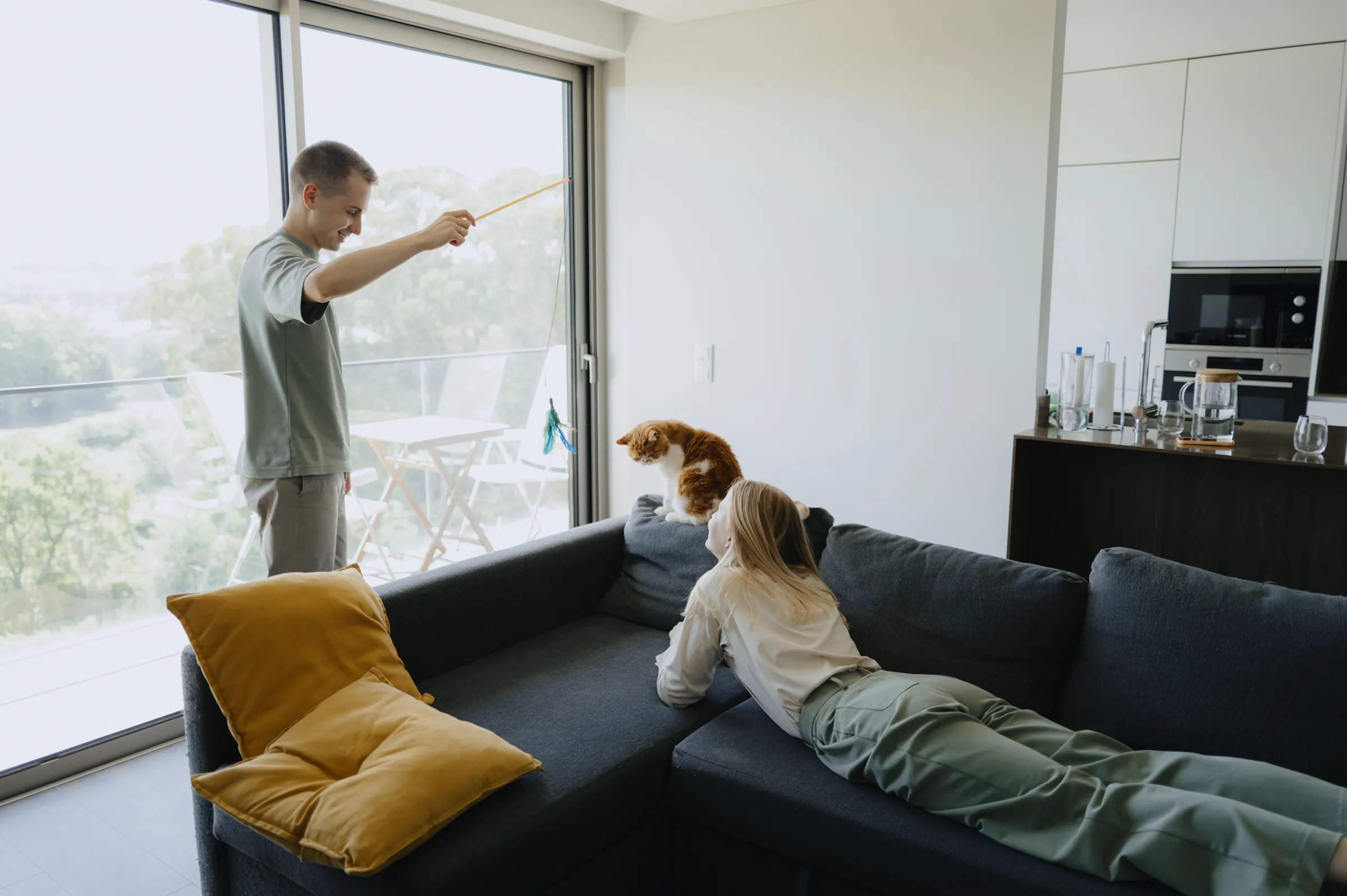 Couple at home with their dog on a dark sofa, soft window light filling the living room.