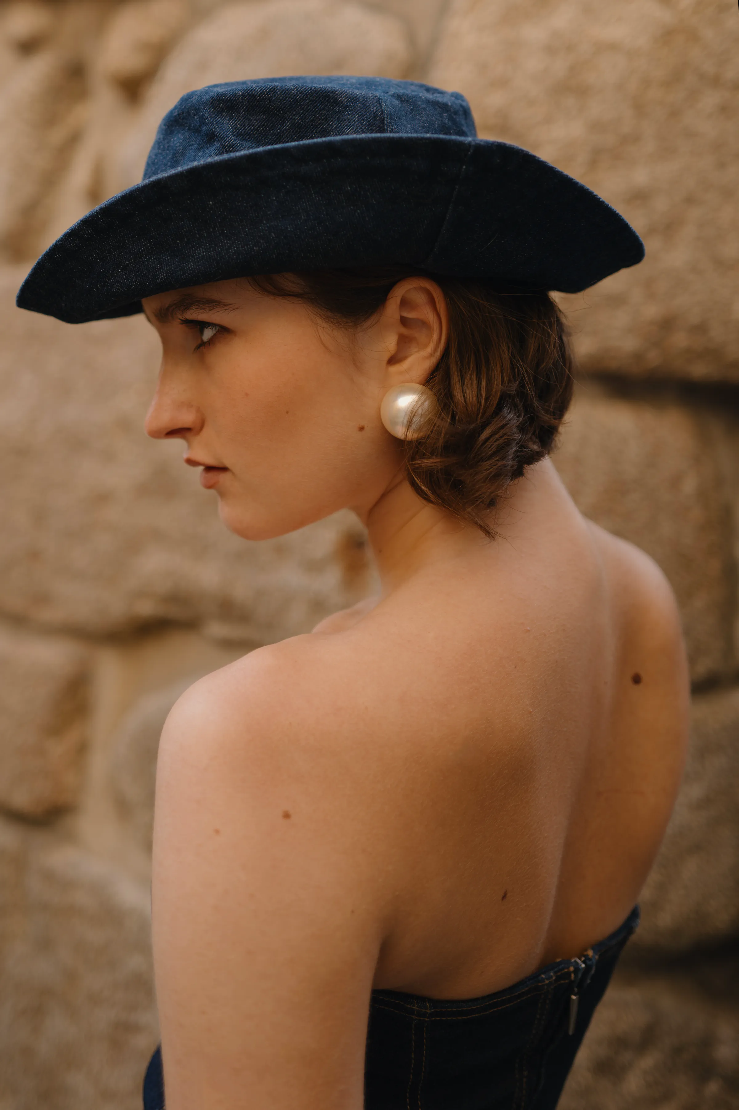 Close portrait of a person in a black wide-brim hat and pearl earring against stone.