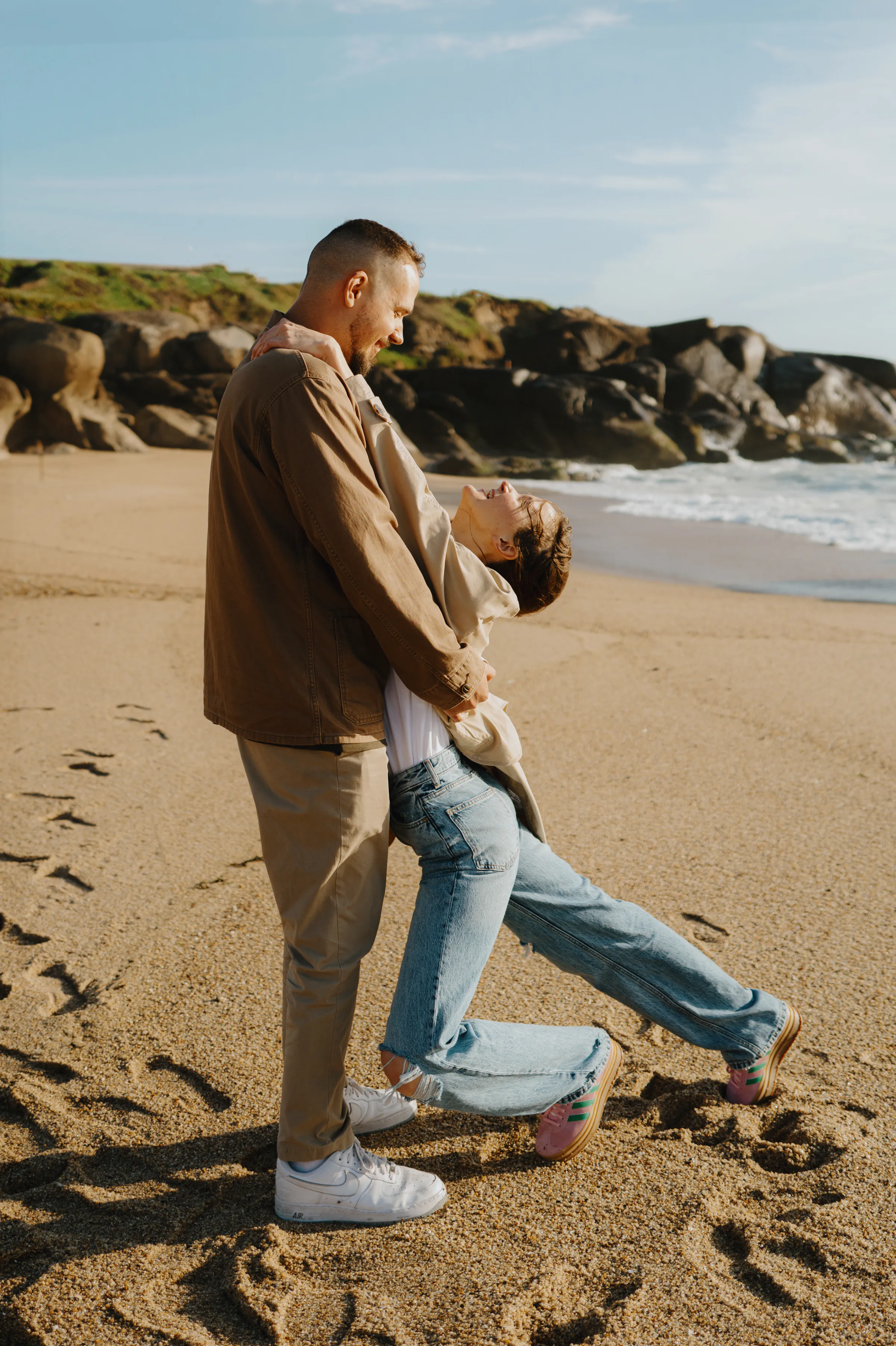 Couple portrait on a sandy seaside beach, standing close near rocks and gentle surf.