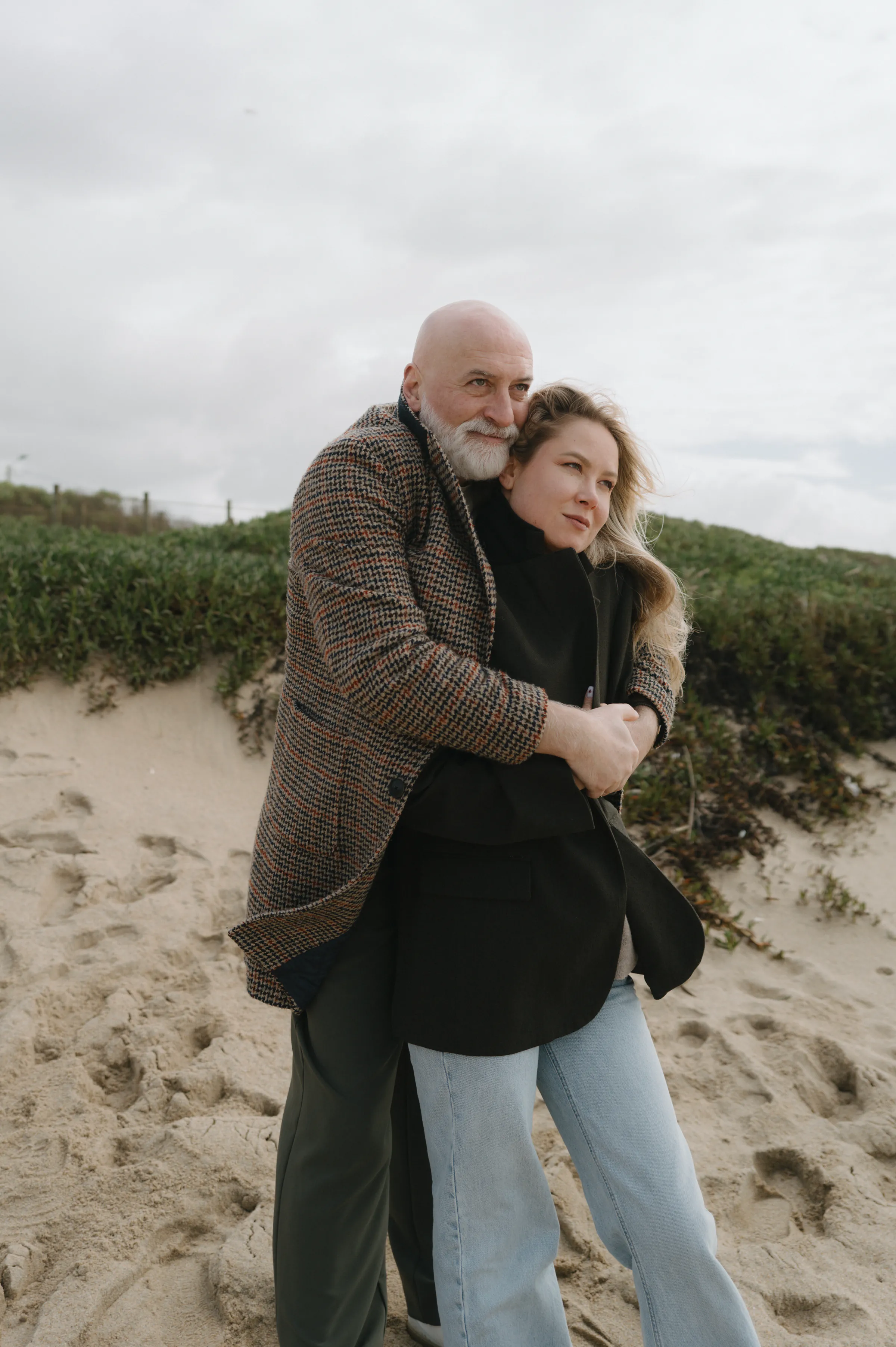 Couple embracing on sandy dunes during an overcast seaside outdoor session.