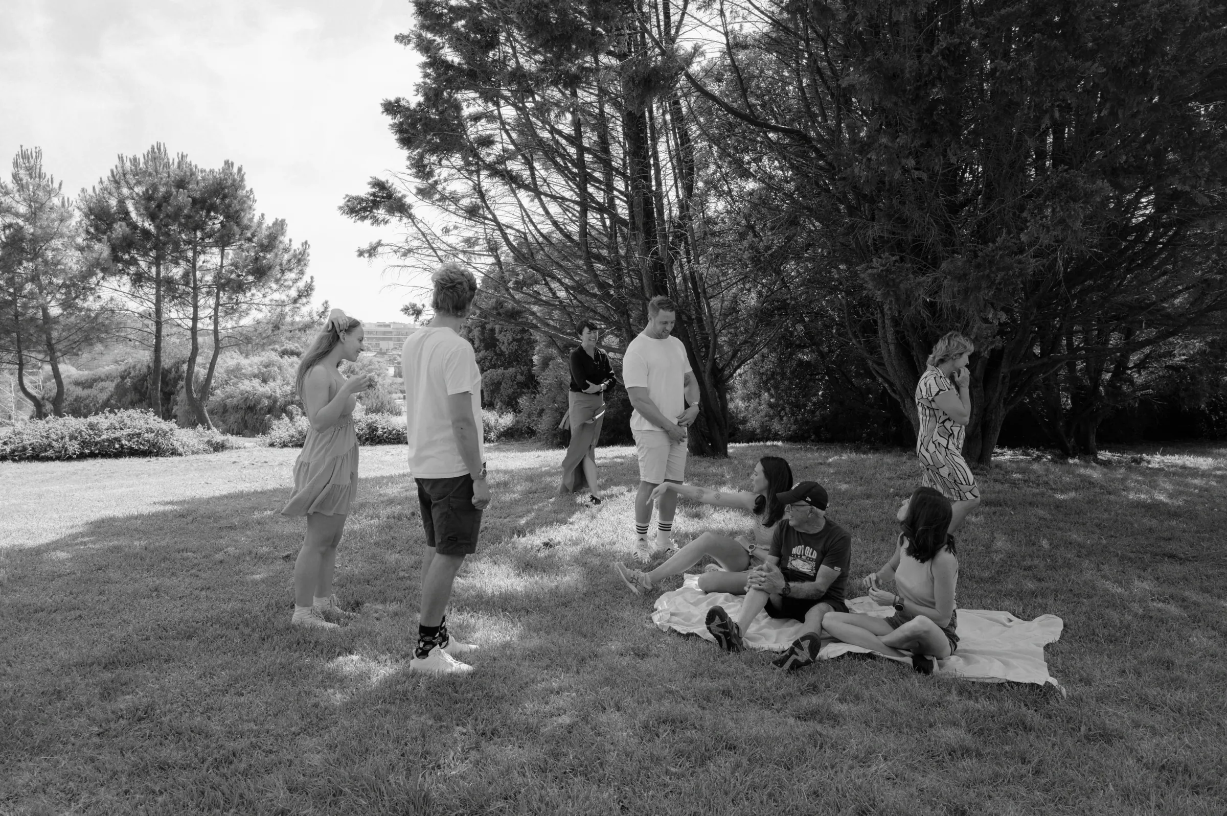 Black-and-white outdoor session with adults and children gathered on grass near trees and a picnic blanket.