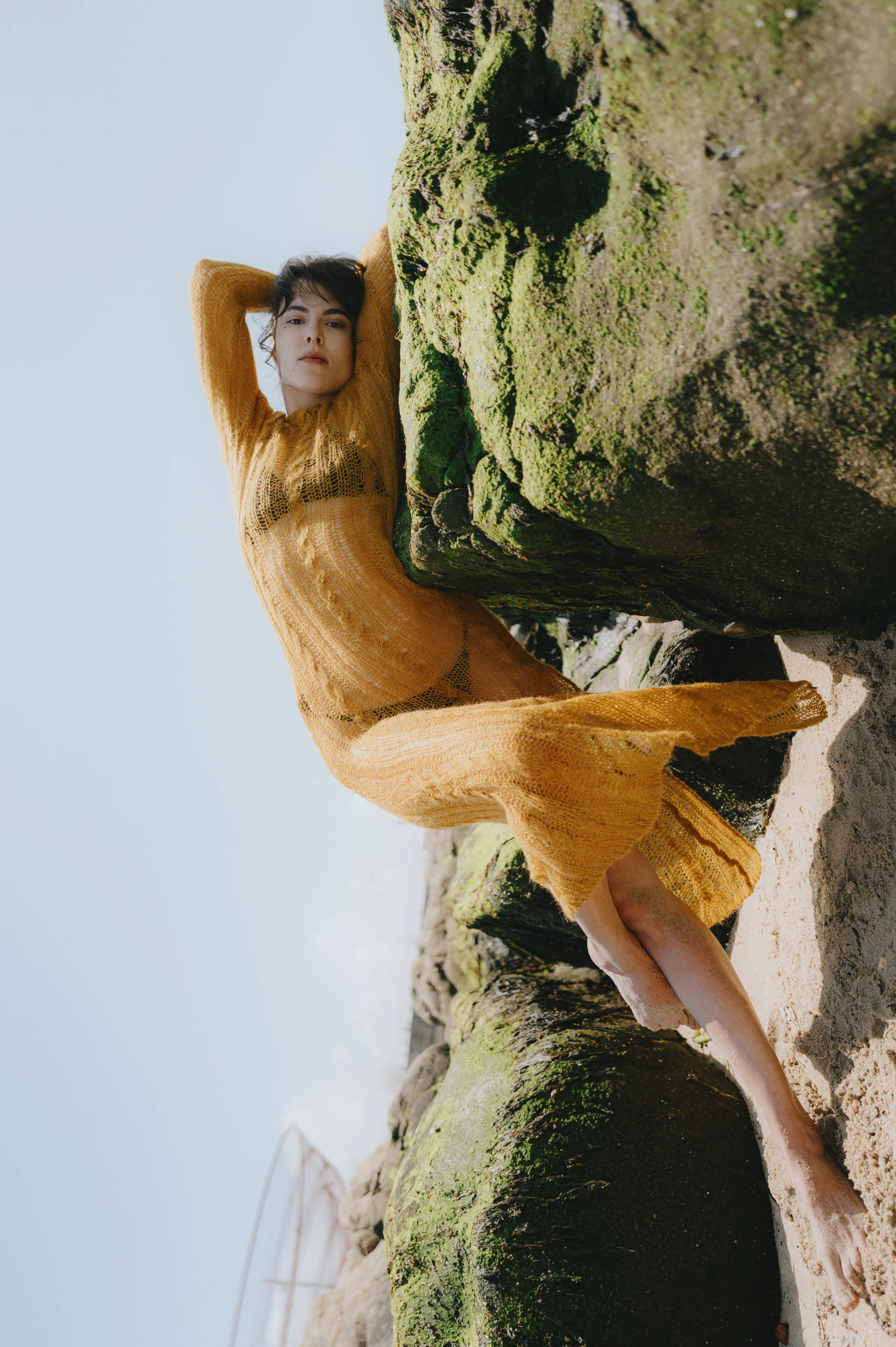 Outdoor seaside portrait of an adult in a mustard knit dress posed against moss-covered rocks in soft light.