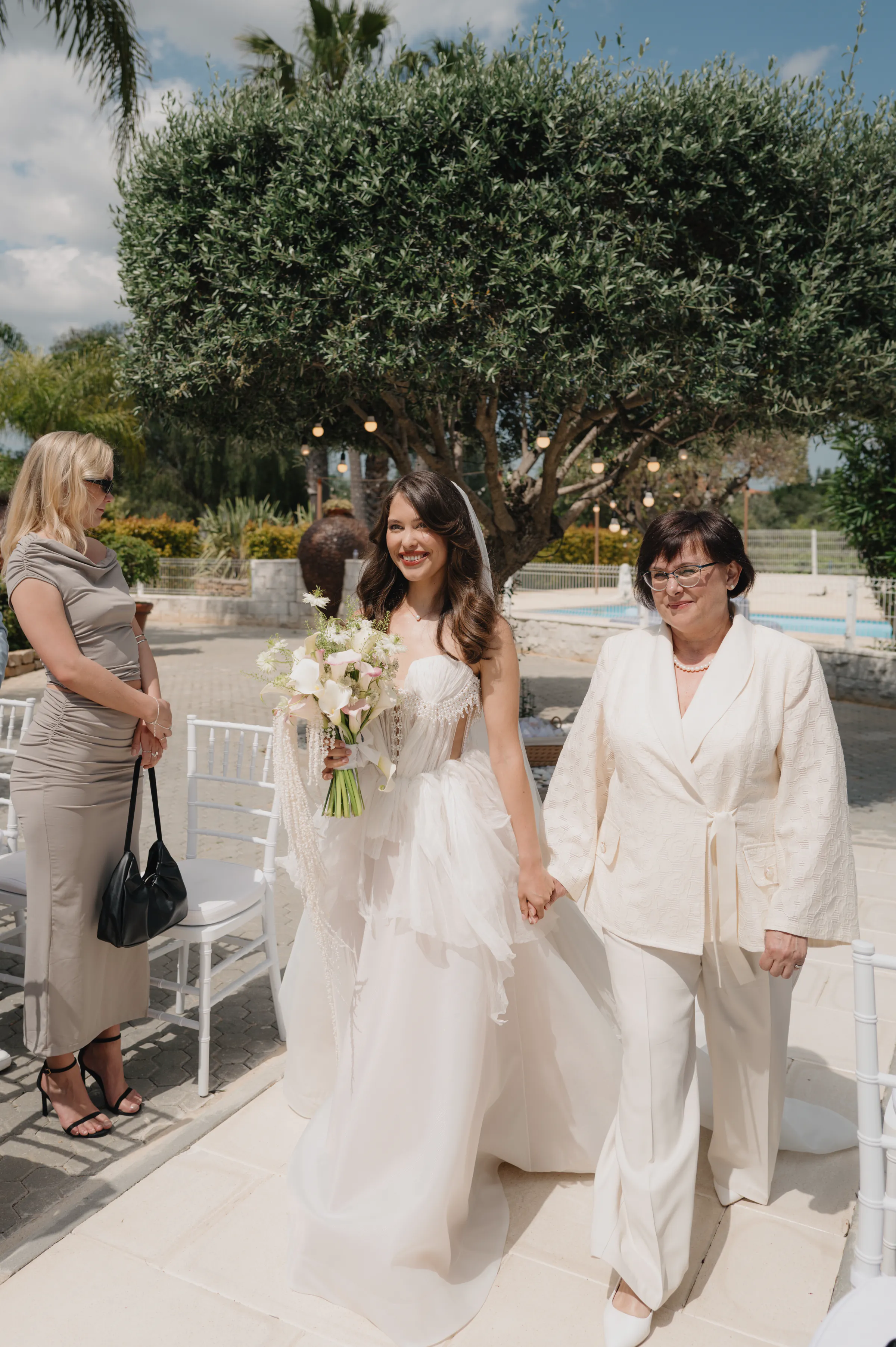 Bride in a flowing gown walks hand in hand with an older adult during an outdoor ceremony.