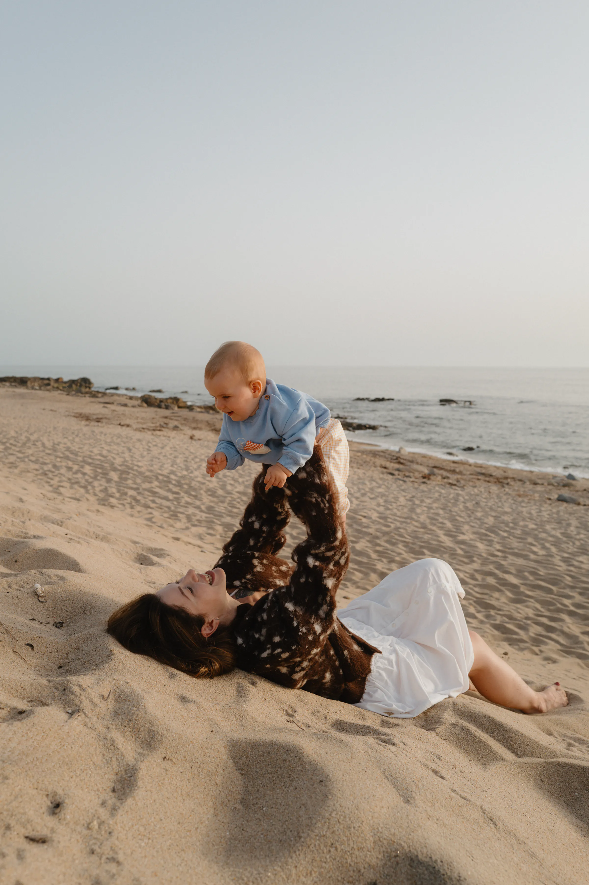 Family seaside portrait with an adult lifting a baby while lying on a sandy beach in soft light