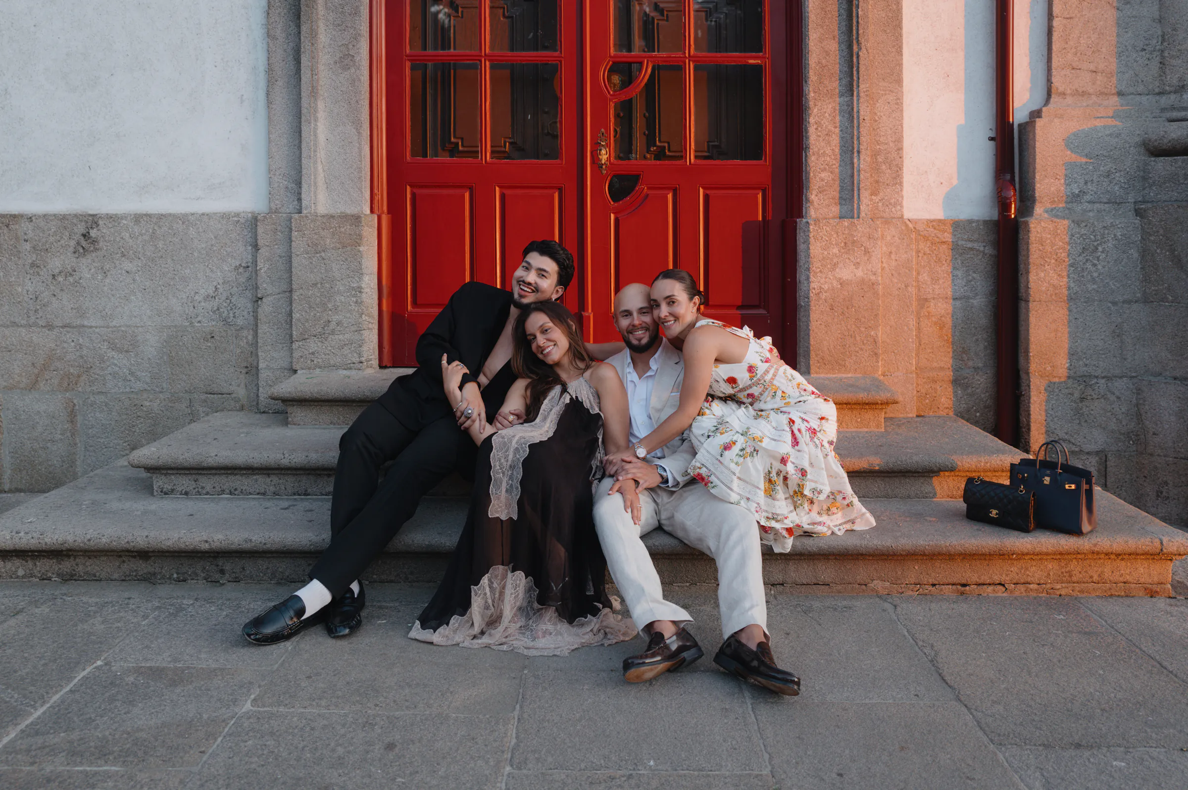 Group portrait of four adults in formal attire seated on stone steps beside tall red doors.