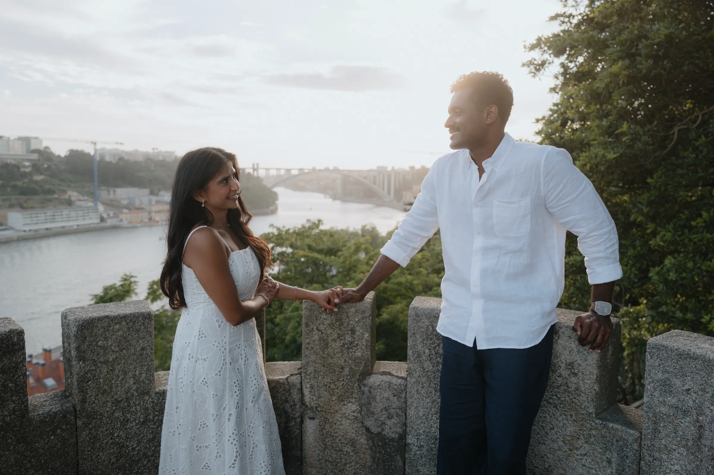 Couple holding hands on a stone overlook above a river during an outdoor city walk in soft light