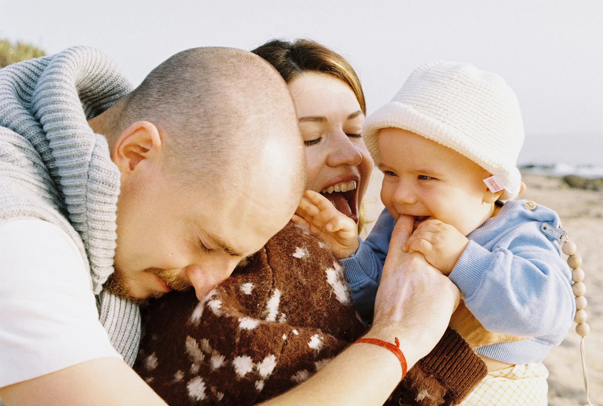 Family close portrait with a baby in a white knit hat during a soft outdoor seaside session
