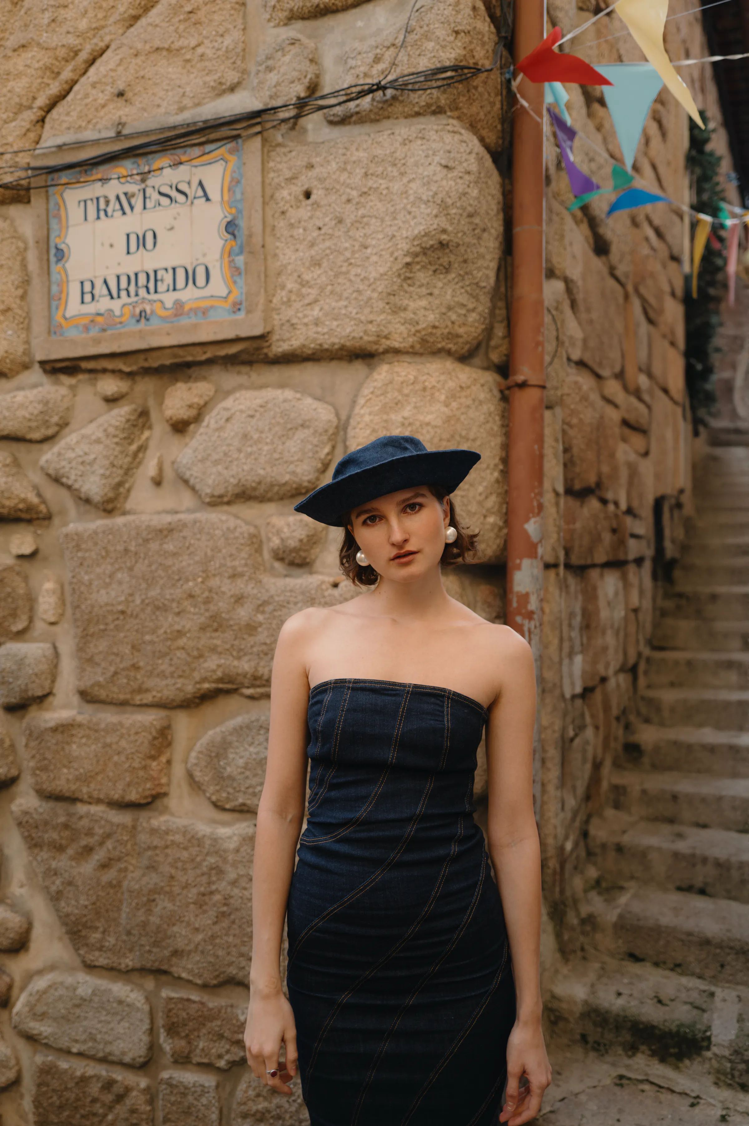 Street portrait of a woman in a dark dress and wide-brim hat beside a textured stone wall.