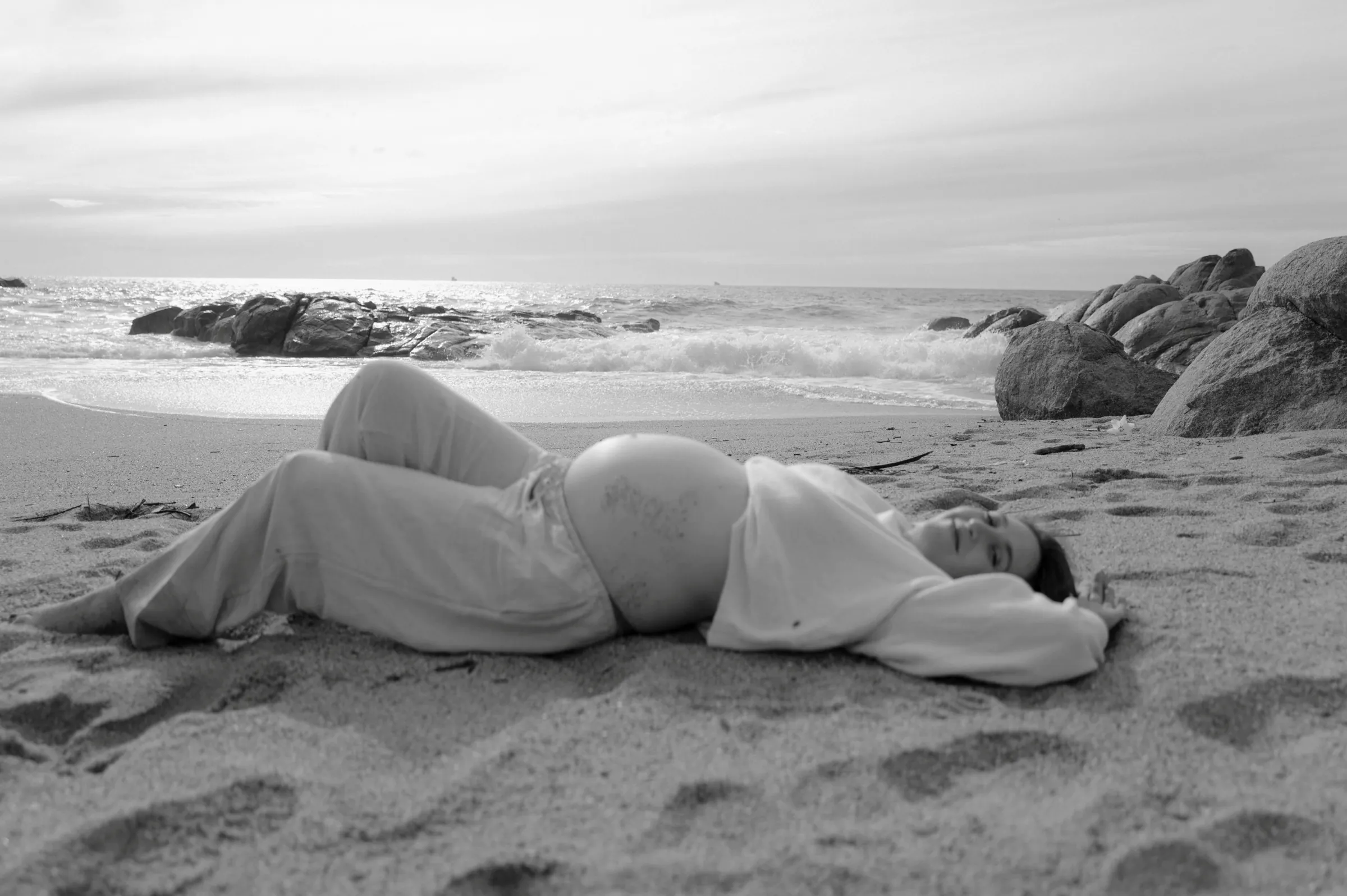 Person reclining on a sandy seaside beach, with rocks and waves in soft black-and-white light.