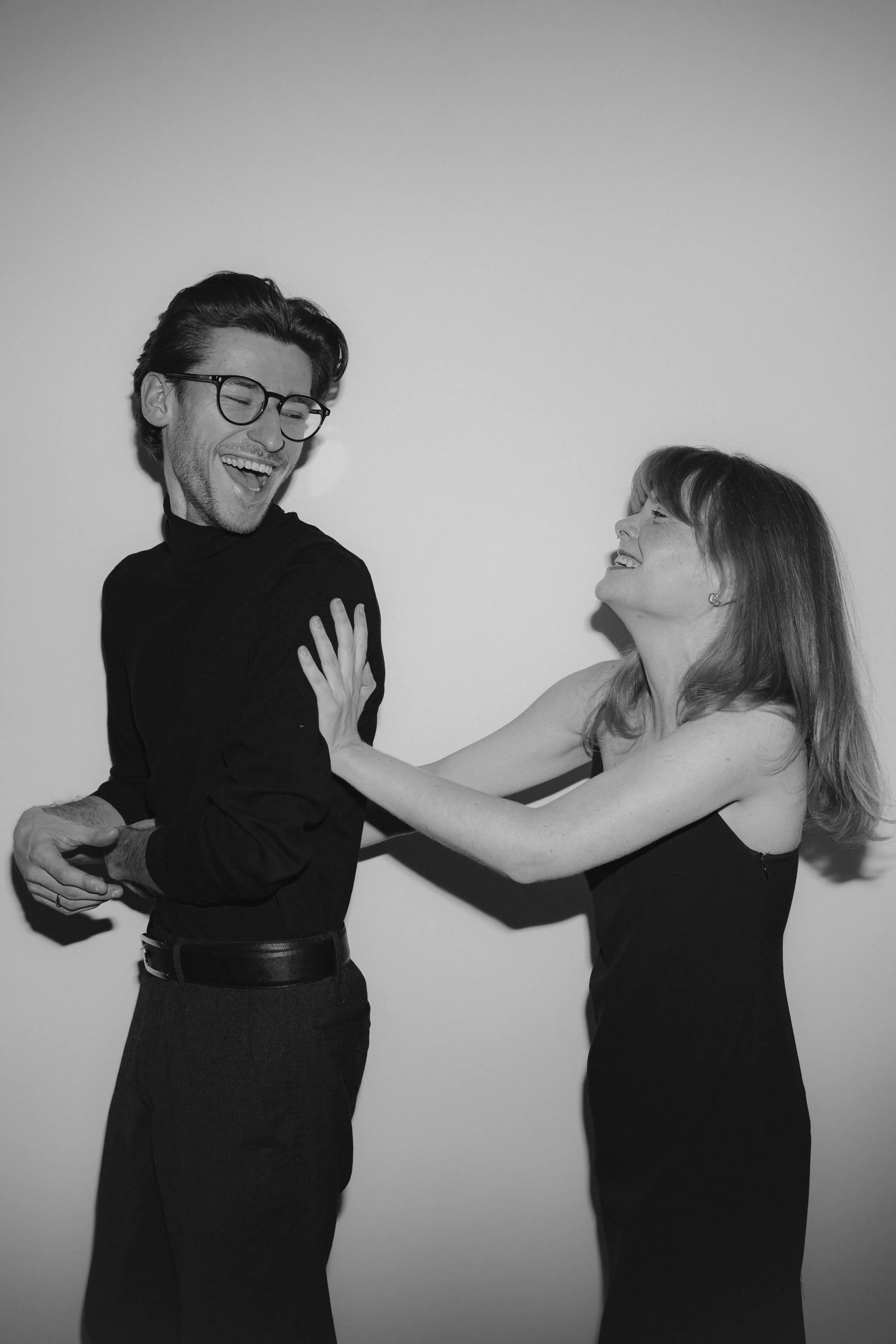 Black-and-white couple portrait with playful posing against a plain studio-style backdrop.