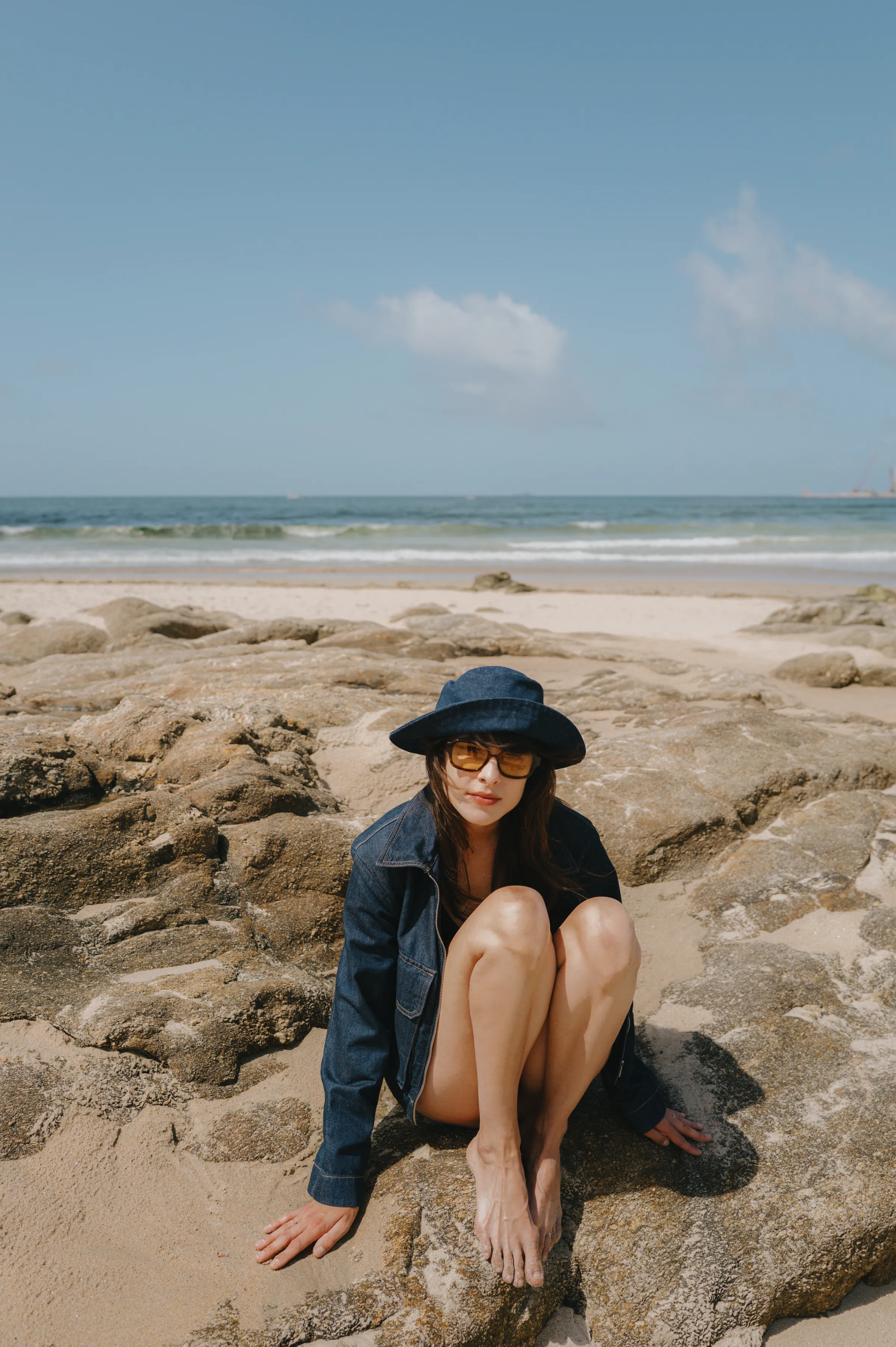Seaside portrait of a person in a blue hat seated on sunlit rocks by the shore.