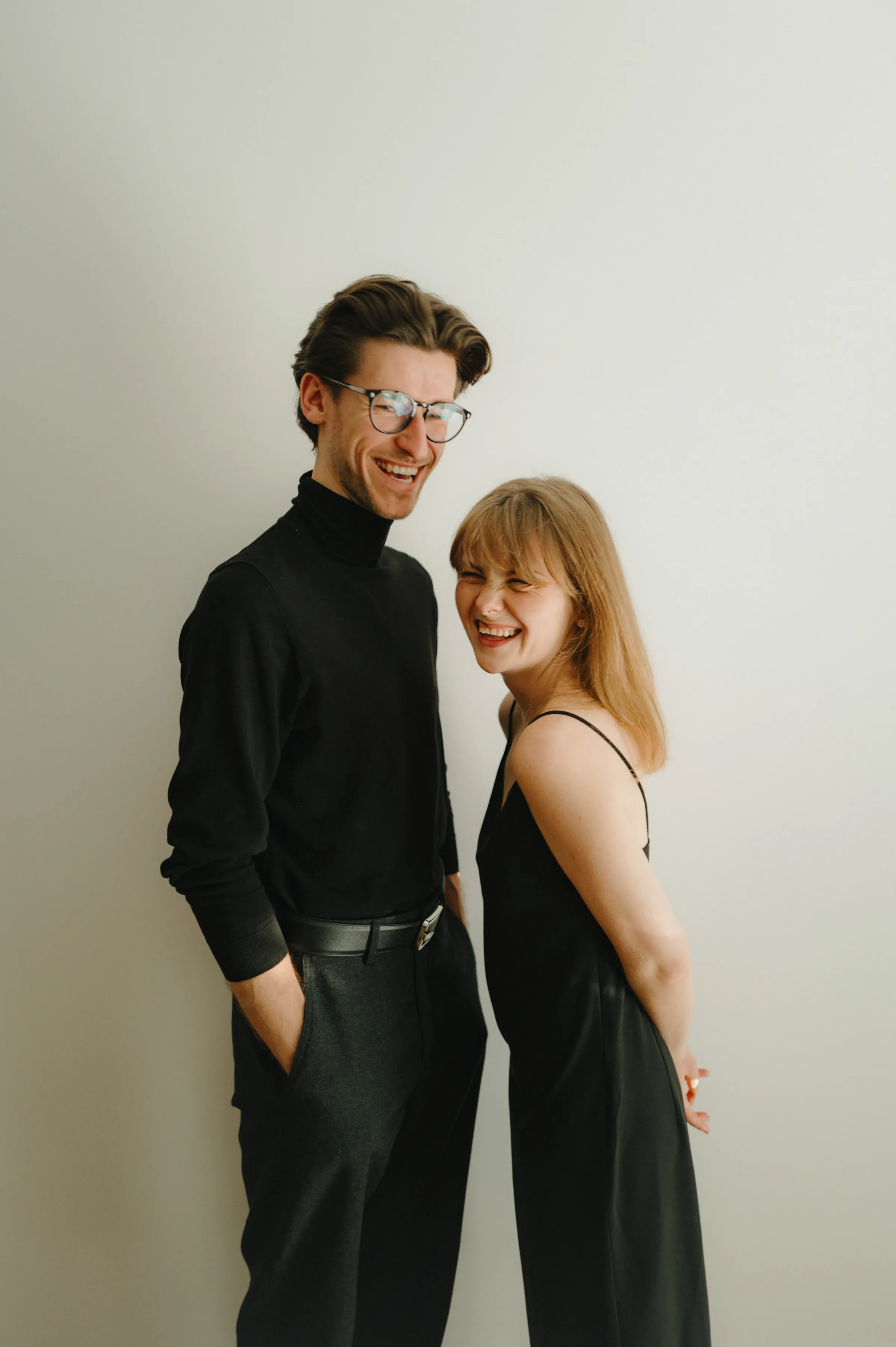Couple in black outfits standing close together for a simple indoor portrait against a plain light wall.