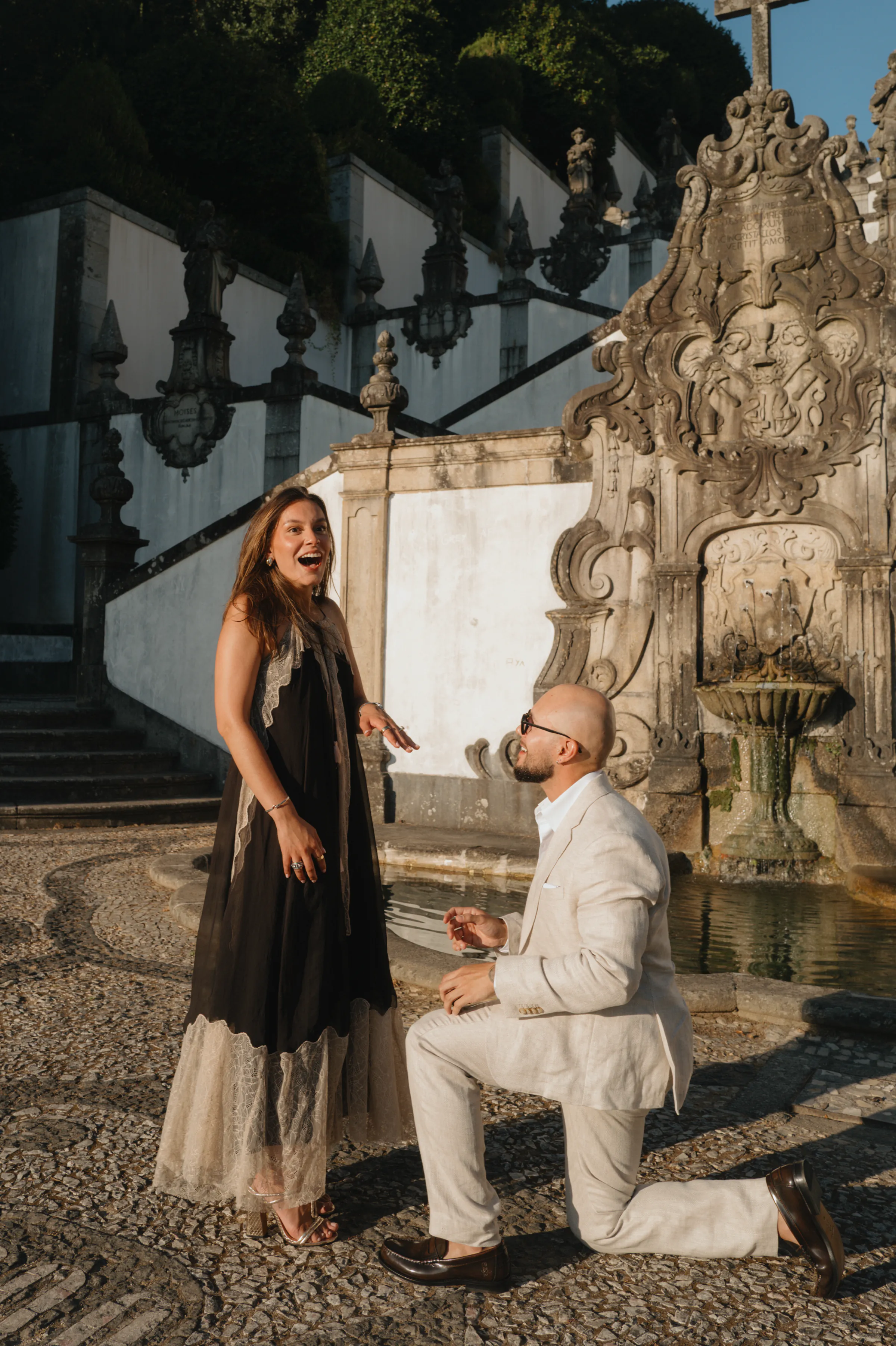 Couple during an outdoor session by an ornate fountain, one kneeling with a ring in warm light.