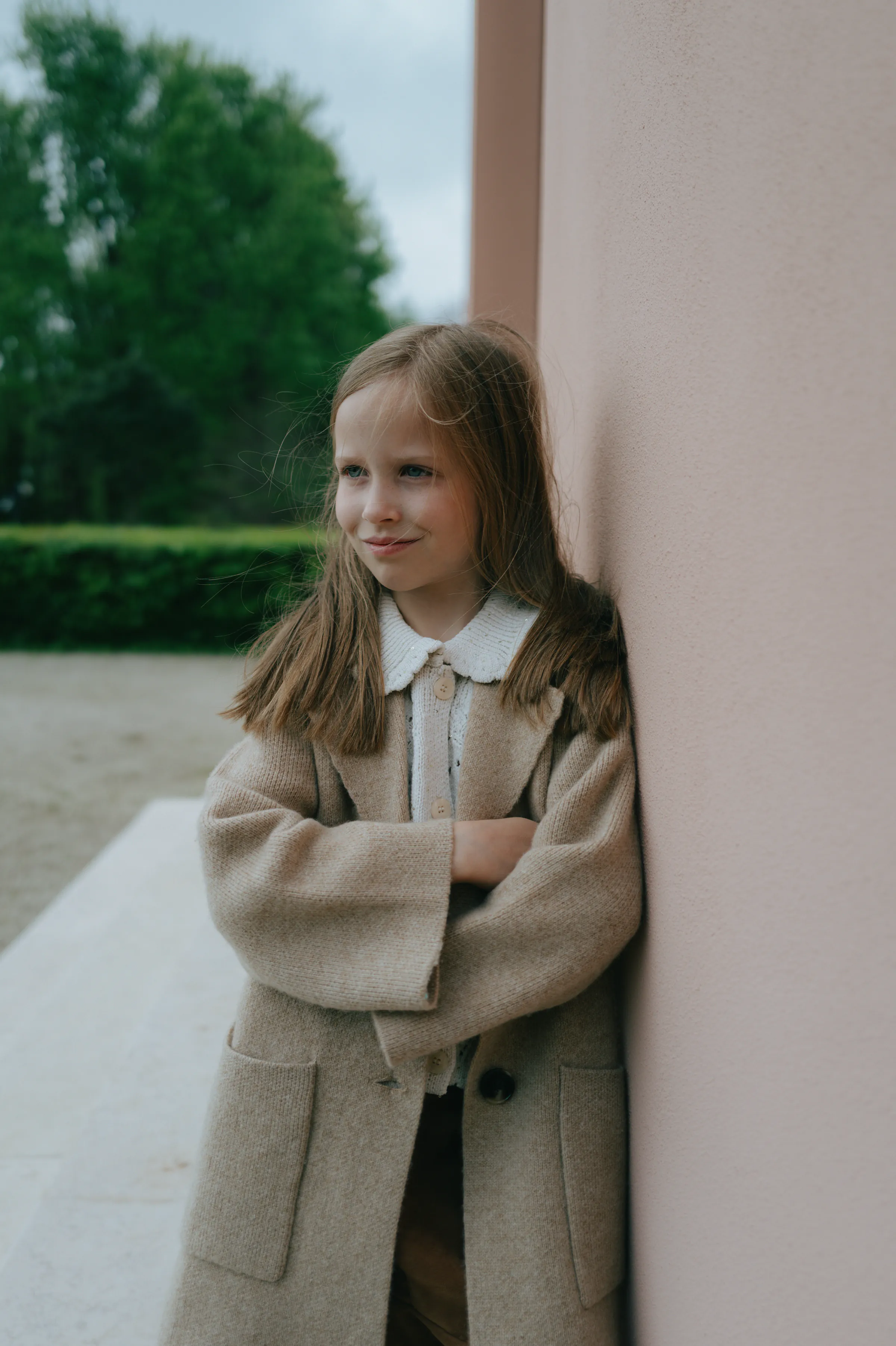 Child portrait leaning against a pink wall outdoors, wearing a beige coat in soft light.