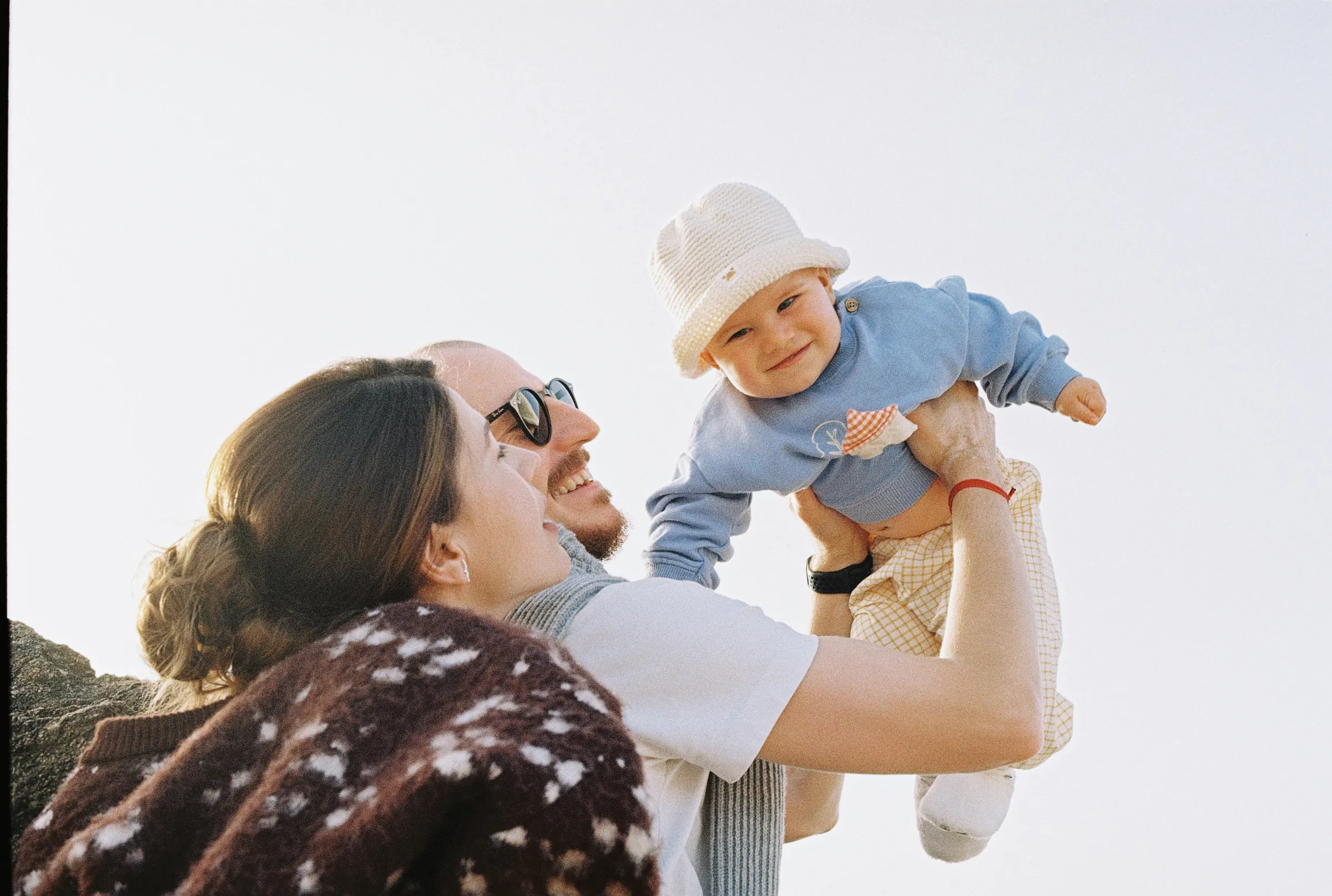 Family outdoor session with two adults lifting a baby in soft light near rocky coastline.