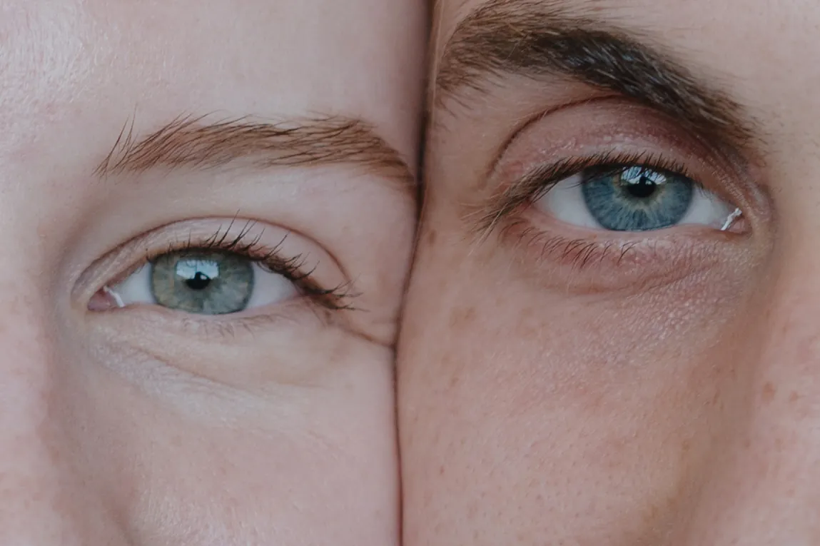 Close portrait of two people pressed cheek to cheek, focused on their eyes in soft light.