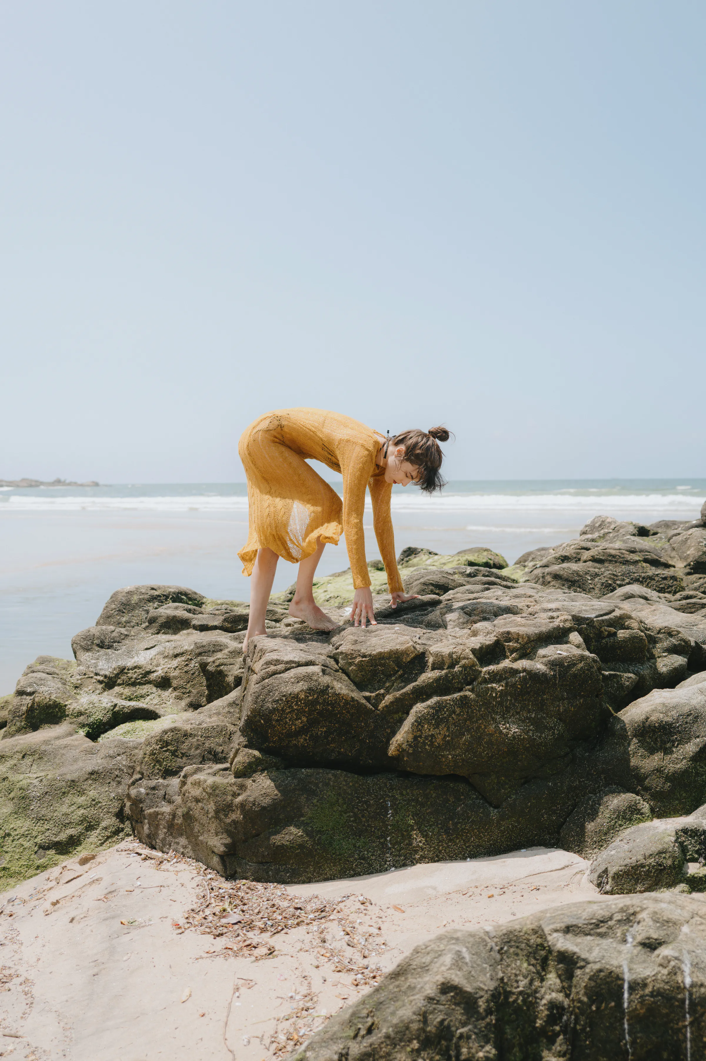 Seaside portrait of a person in a yellow dress bending over sunlit rocks near the shore.