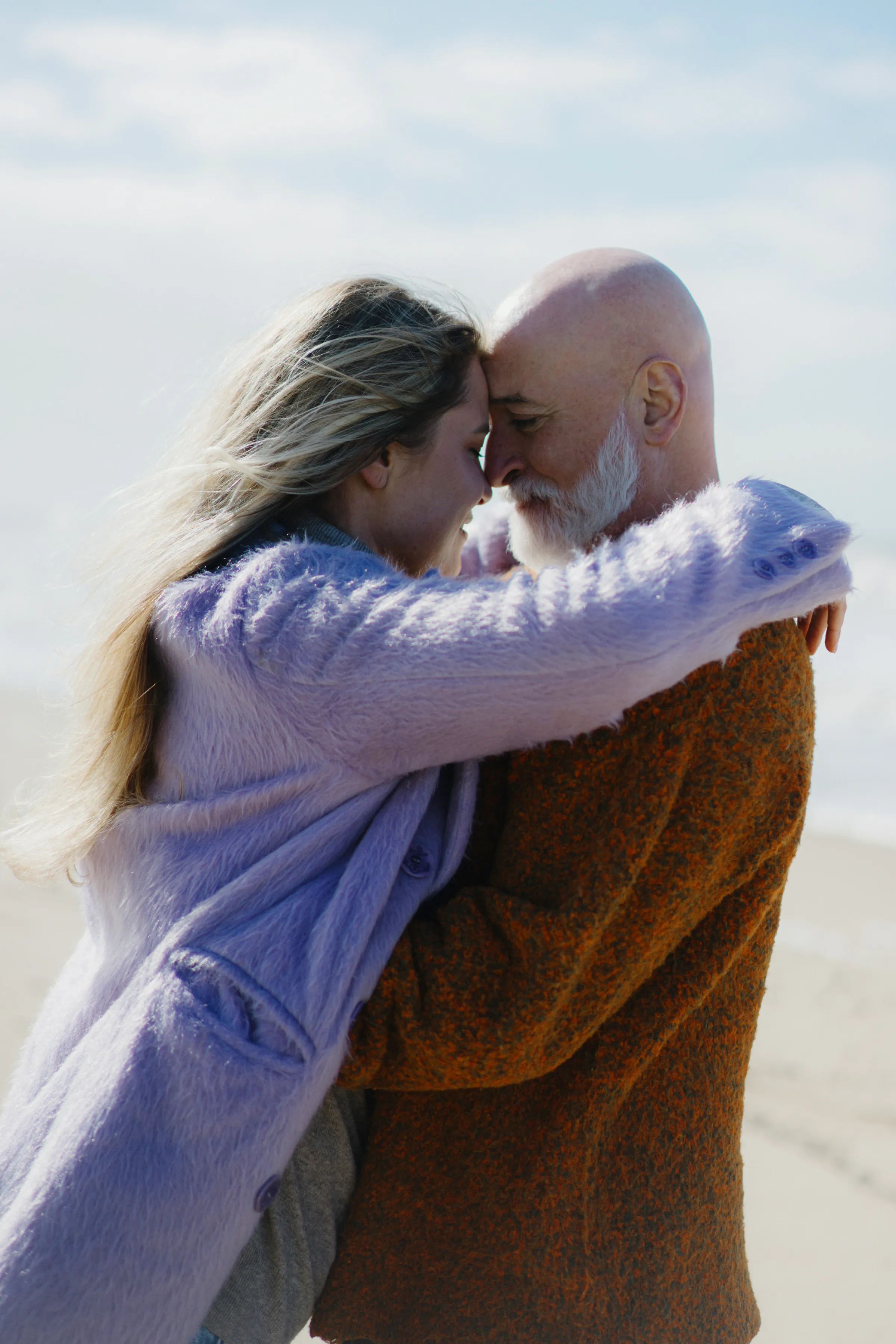 Couple embracing on a breezy seaside session, foreheads touching in soft light.