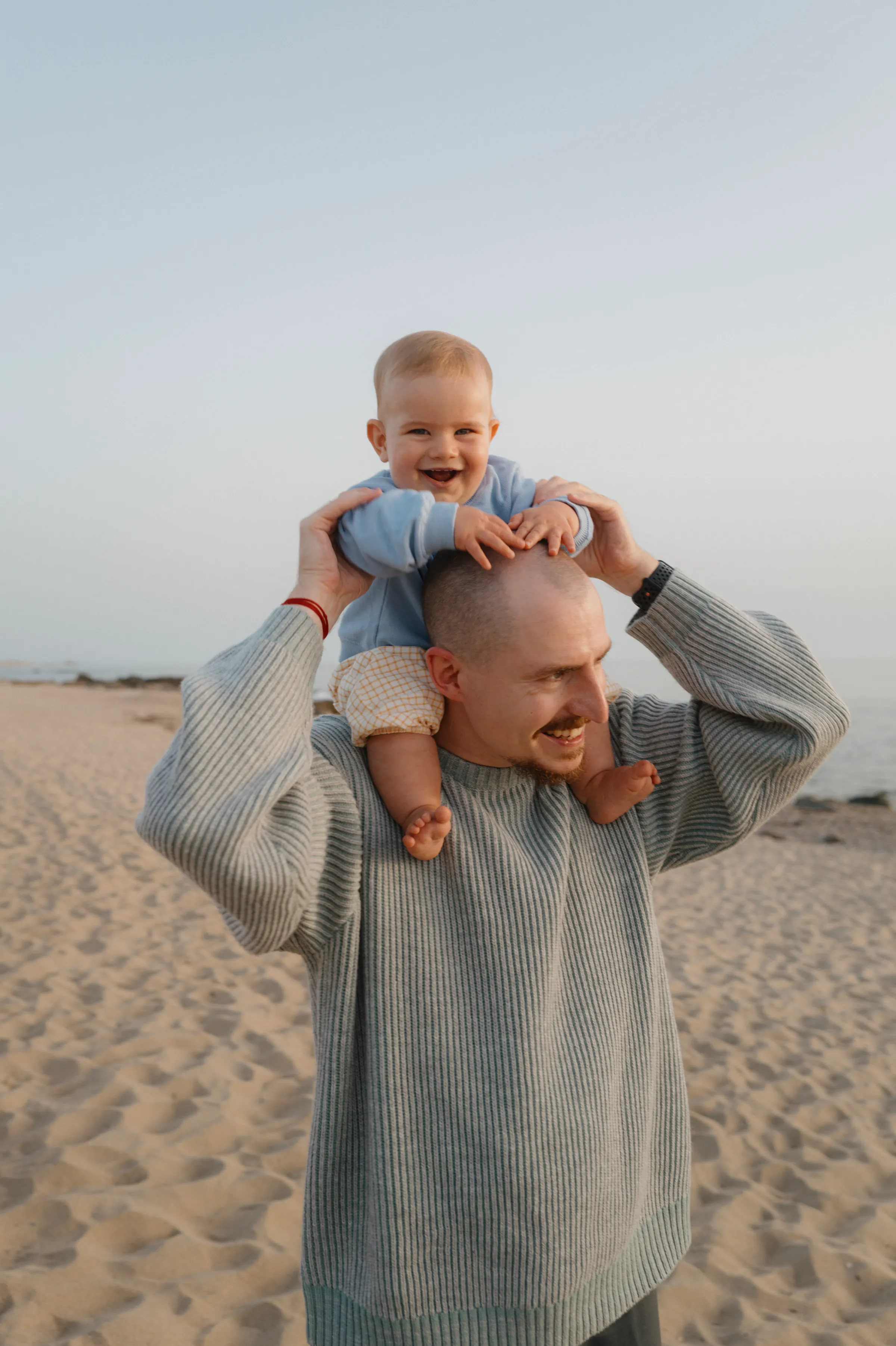 Seaside family portrait of an adult carrying a baby on his shoulders in soft light