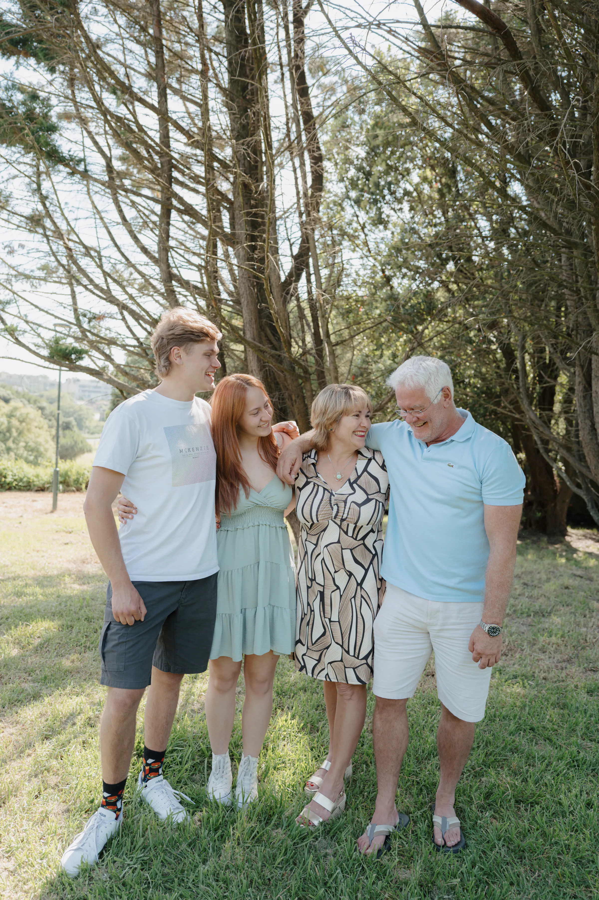 Four-person outdoor portrait standing arm in arm under tall trees in soft light.