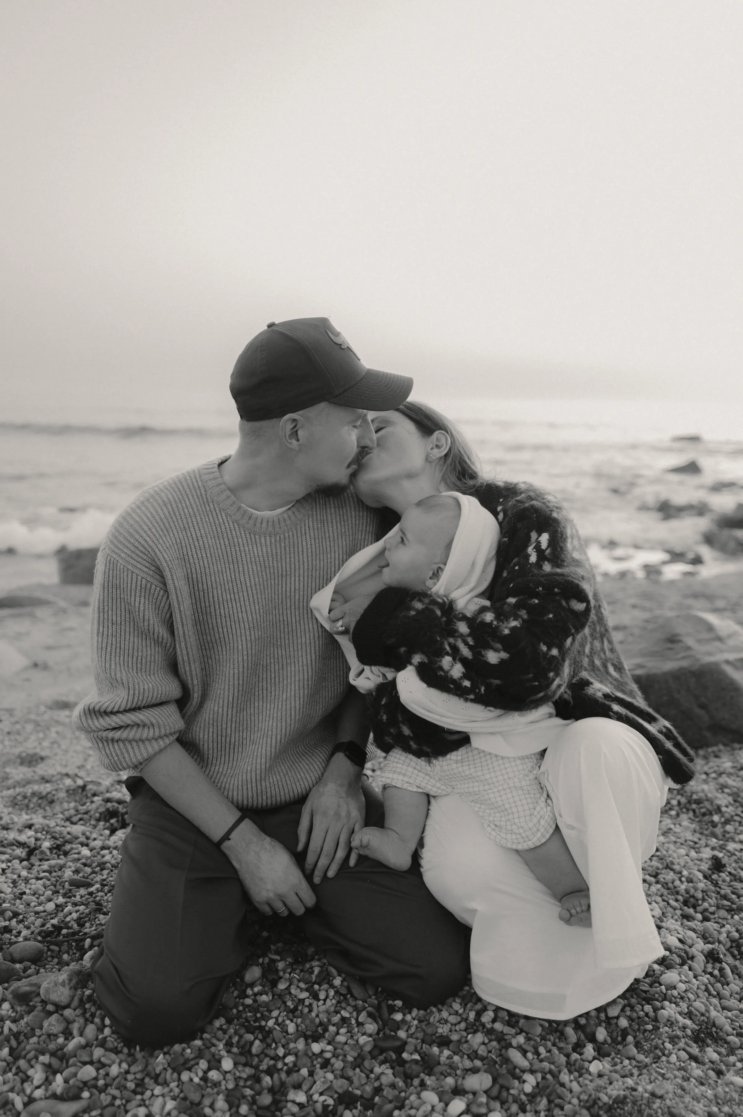 Black-and-white seaside family portrait with a couple kissing while holding a baby on a rocky shore.