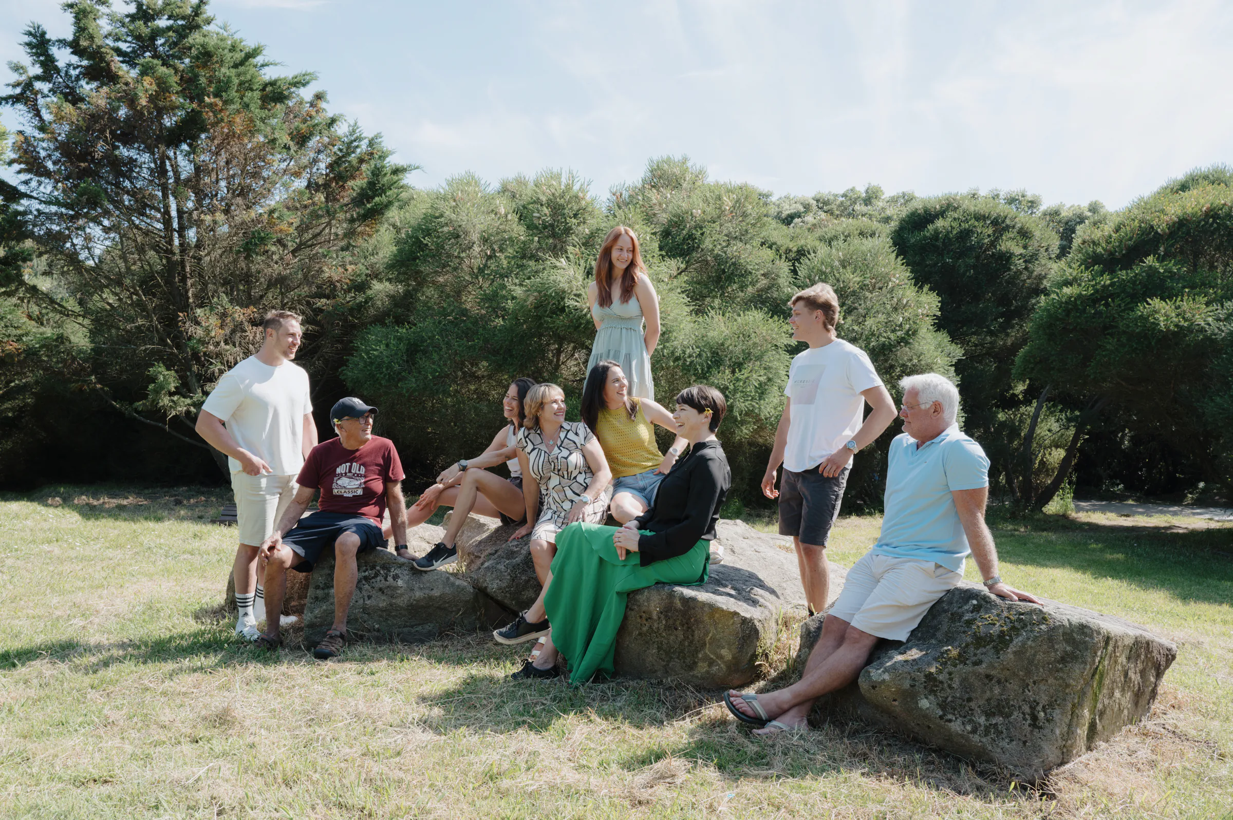 Family outdoor session gathered on large rocks in a sunny park with trees.