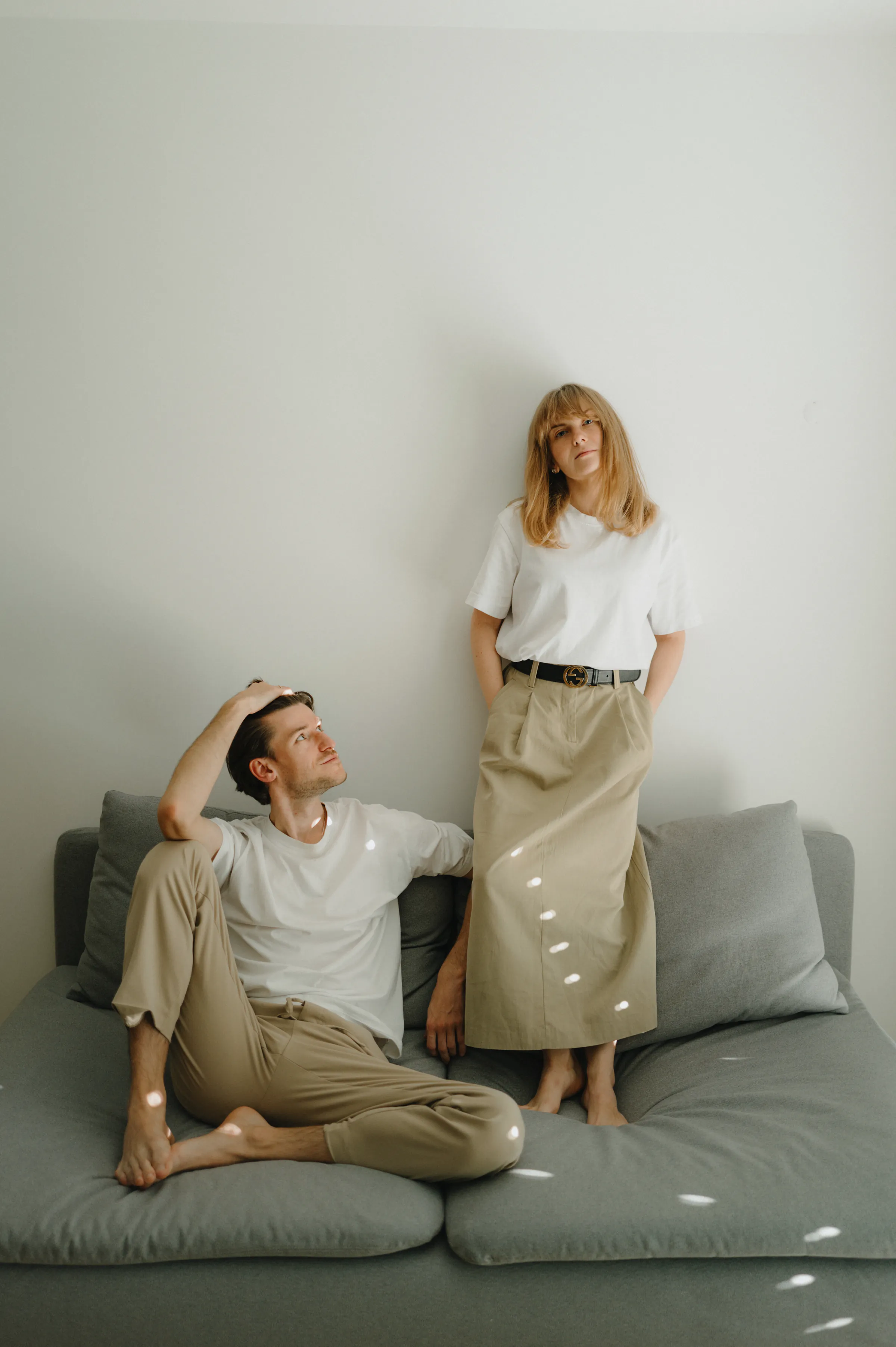 Couple portrait in a minimal home session, one seated on a gray sofa and one standing in soft light.
