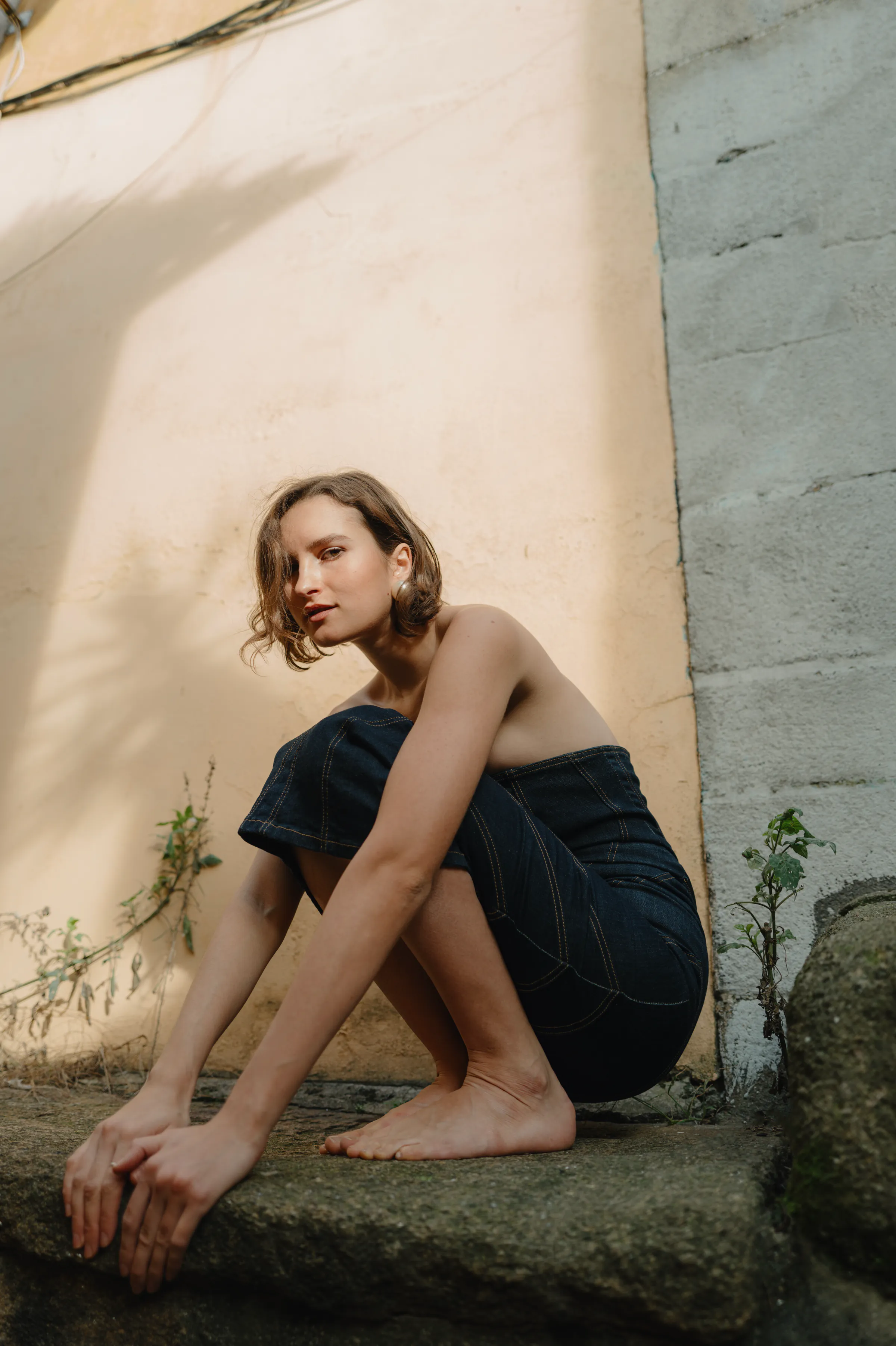 Outdoor portrait of a seated person in a dark dress beside a textured wall in soft light.
