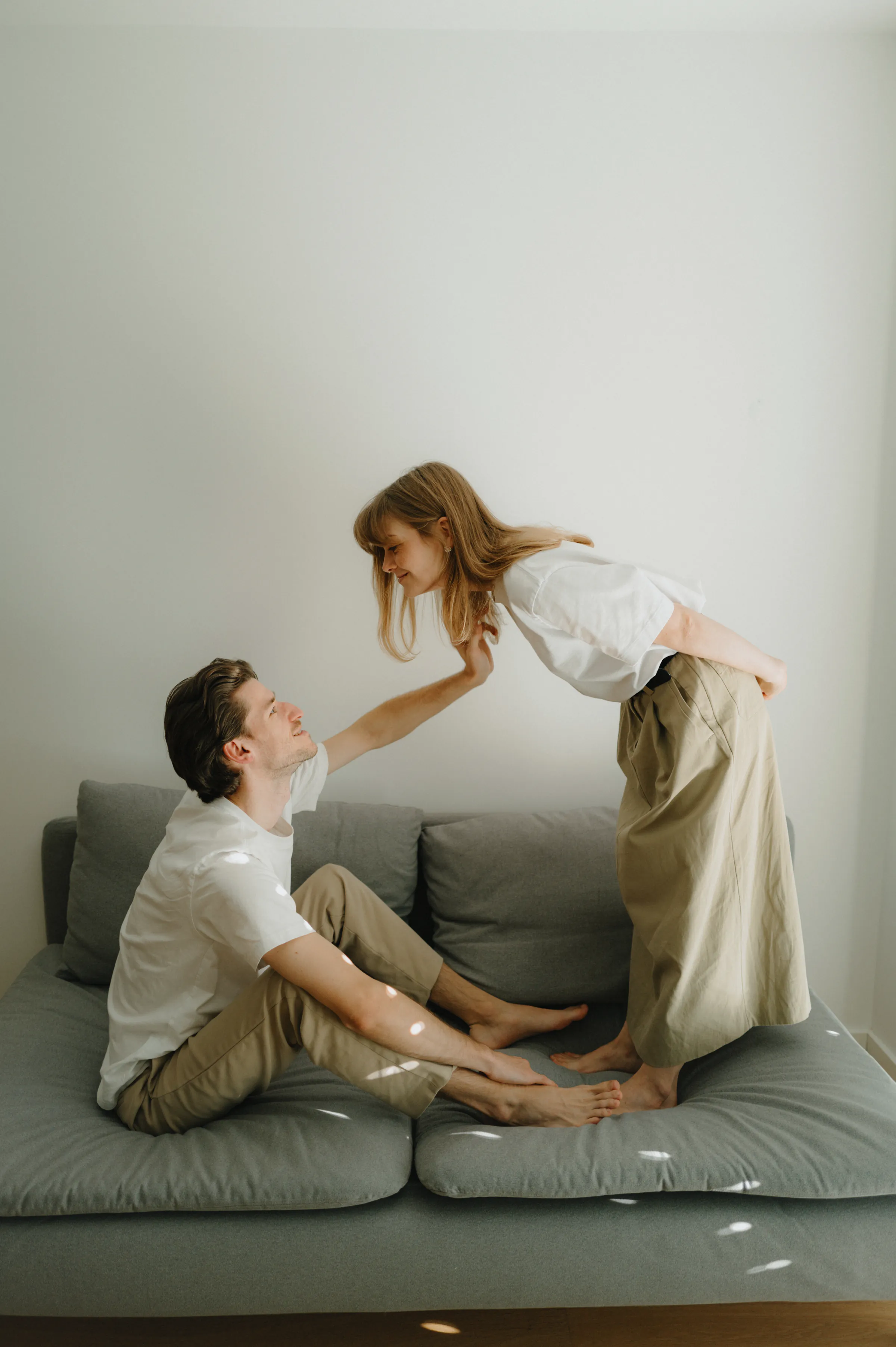 Couple in a home session on a gray sofa, one seated while the other leans close in soft light.