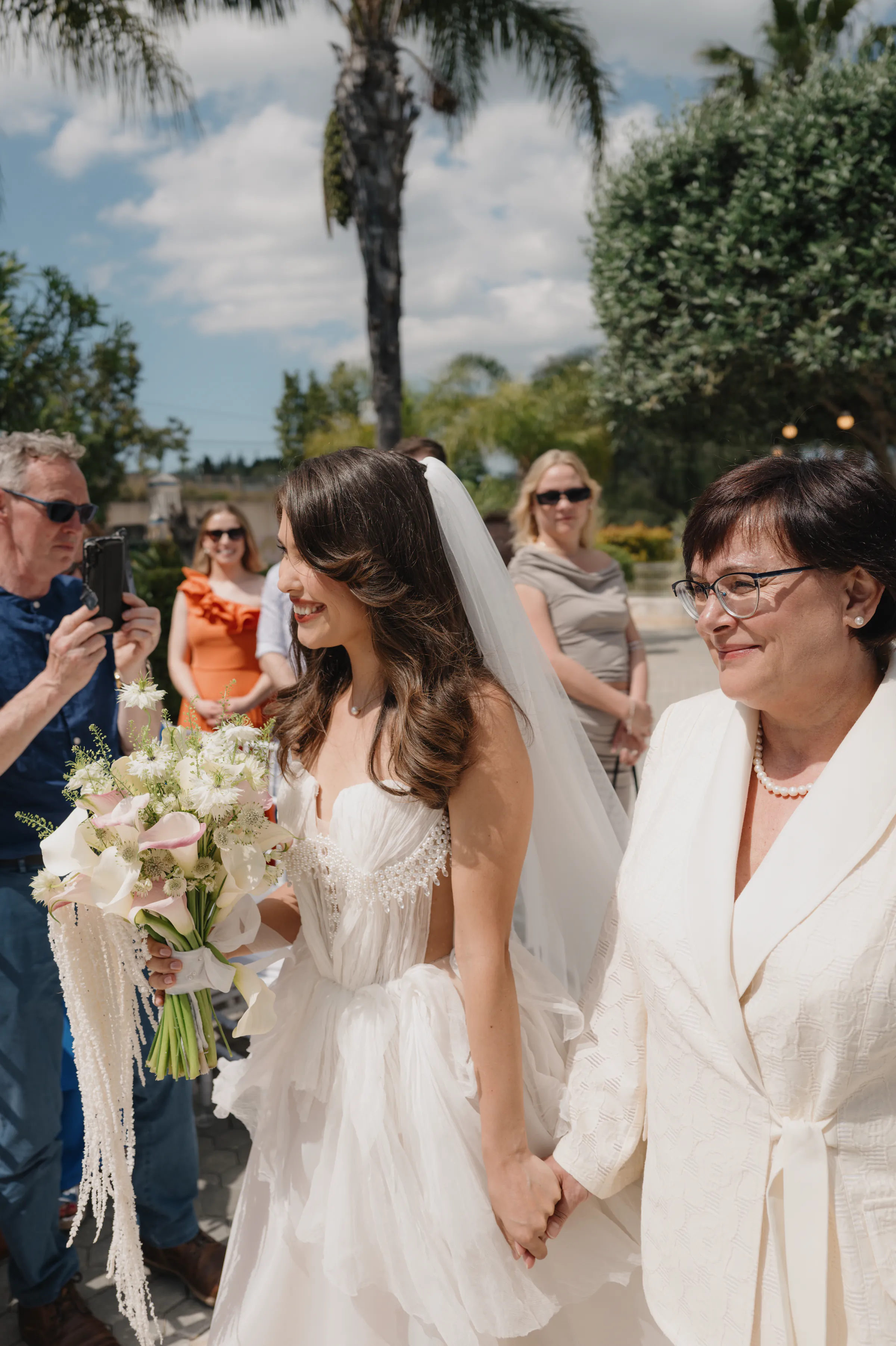 Bride holding a bouquet and a woman’s hand during an outdoor wedding ceremony with guests nearby