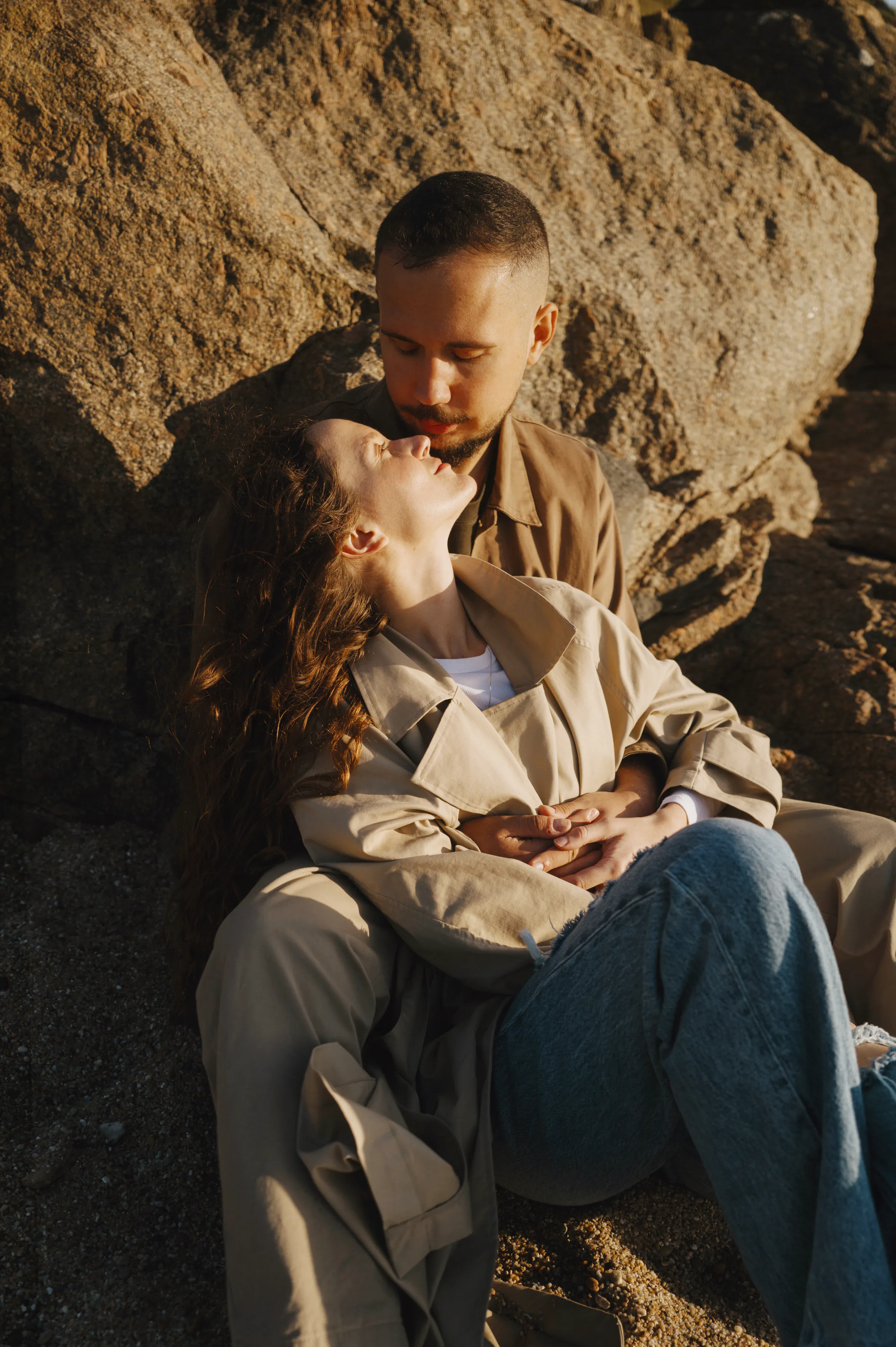 Couple close portrait seated against sunlit rocks during an outdoor session by the shore.