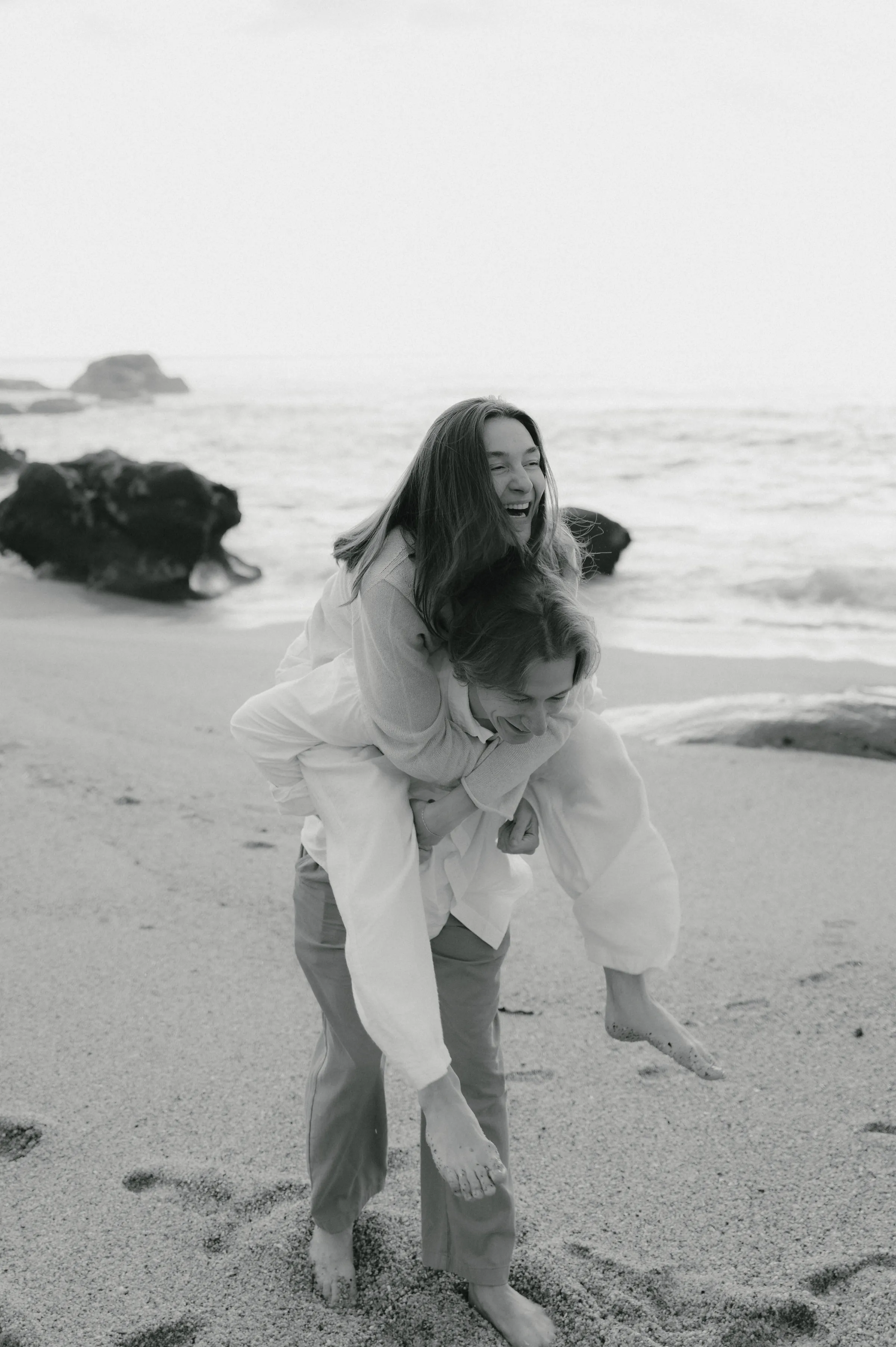 Black-and-white seaside couple portrait during a playful piggyback moment on a sandy beach near waves and rocks.