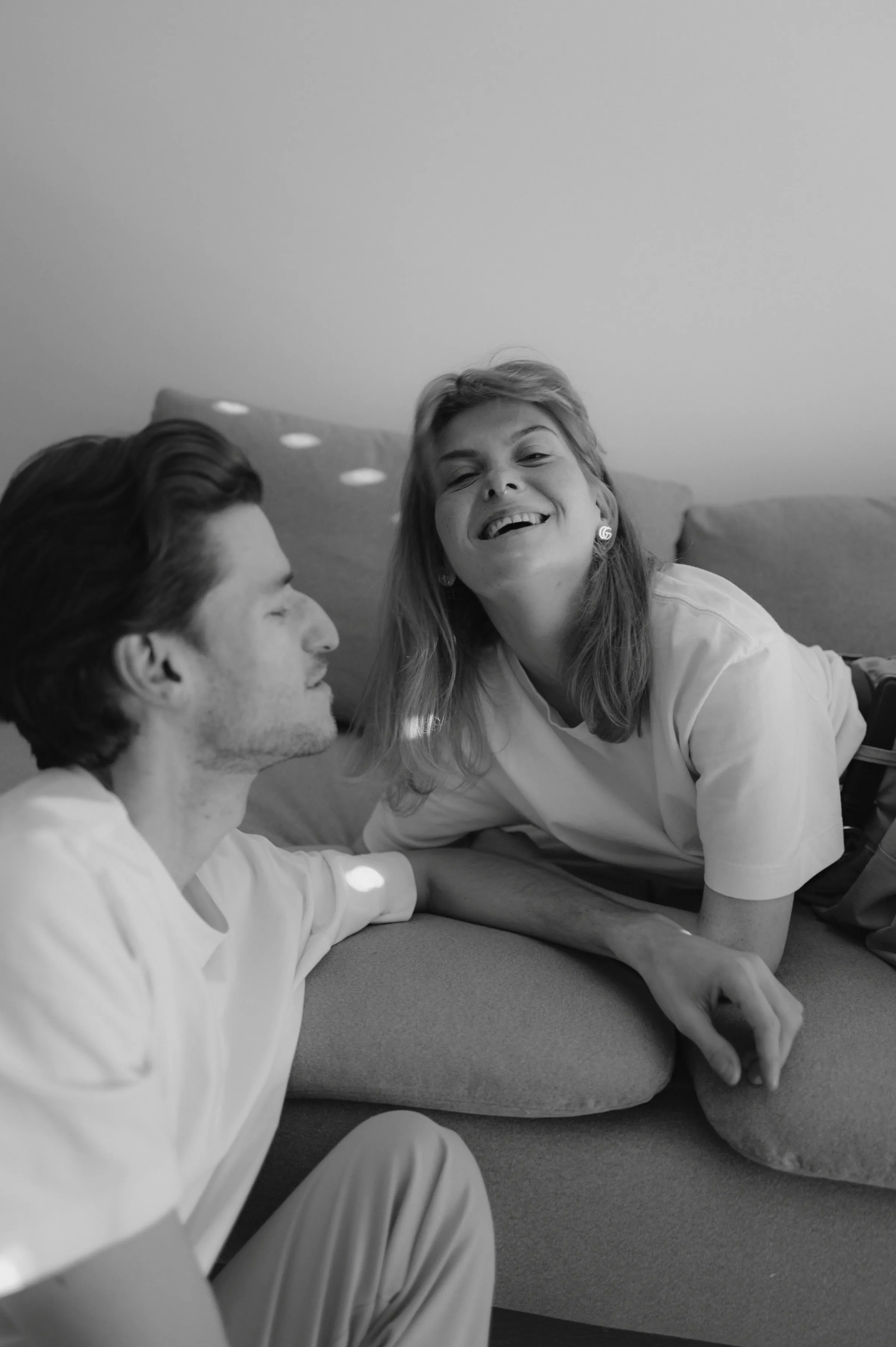 Black-and-white home session portrait of a couple sitting close together on a sofa in soft light.