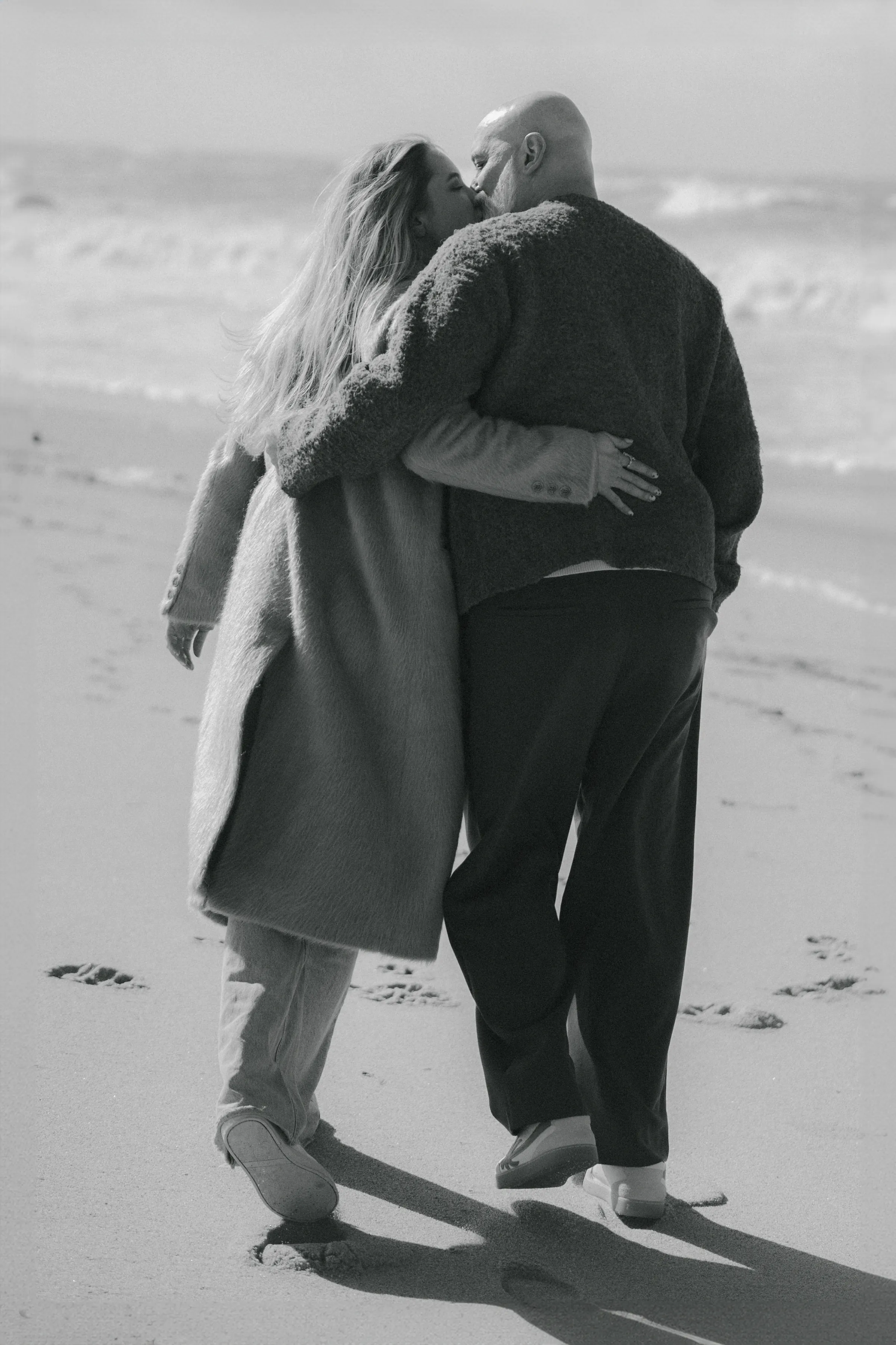 Couple walking arm in arm along a seaside beach in a black-and-white outdoor session.