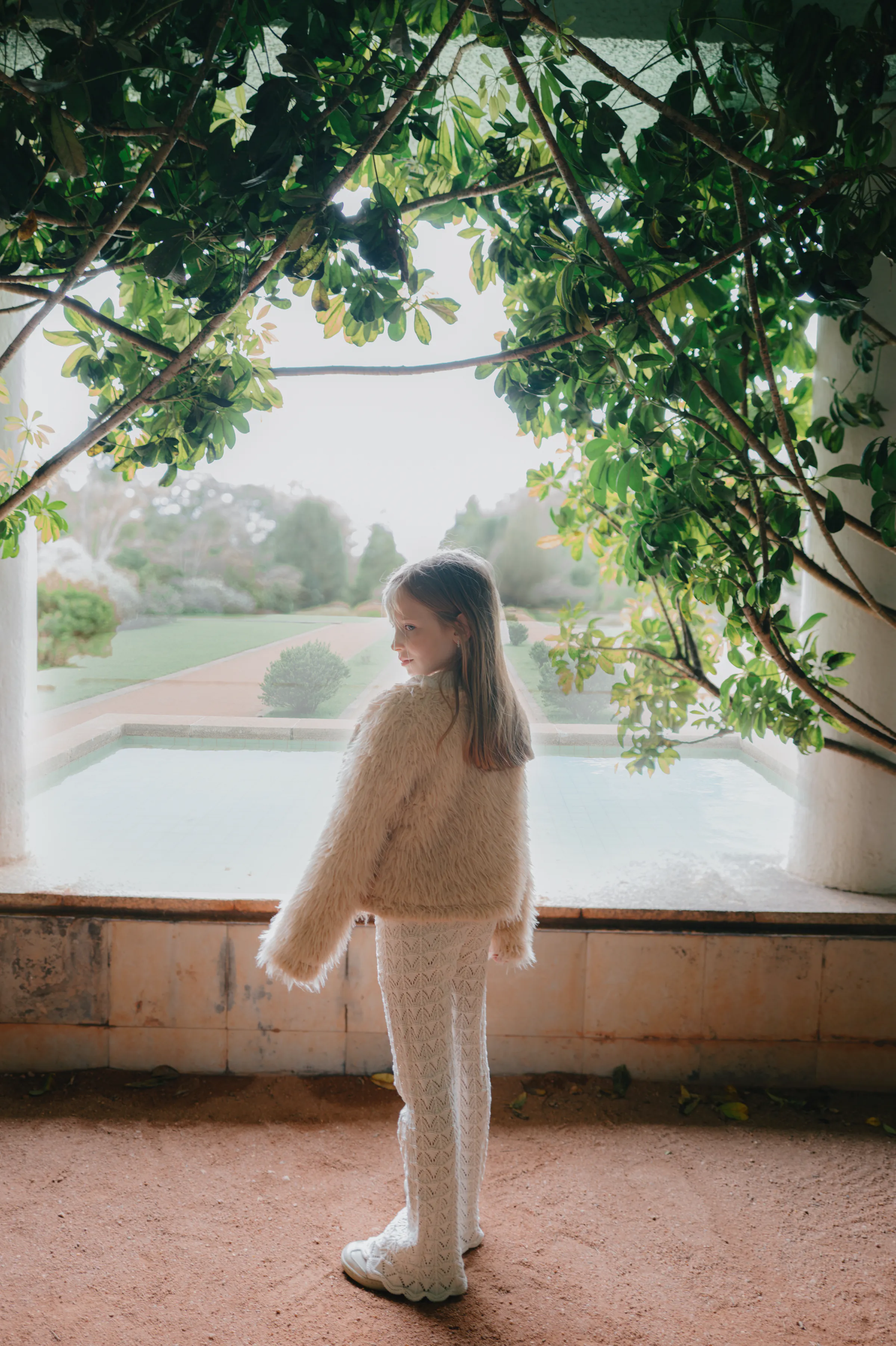 Child portrait in soft light, standing beneath leafy vines beside a garden pool.