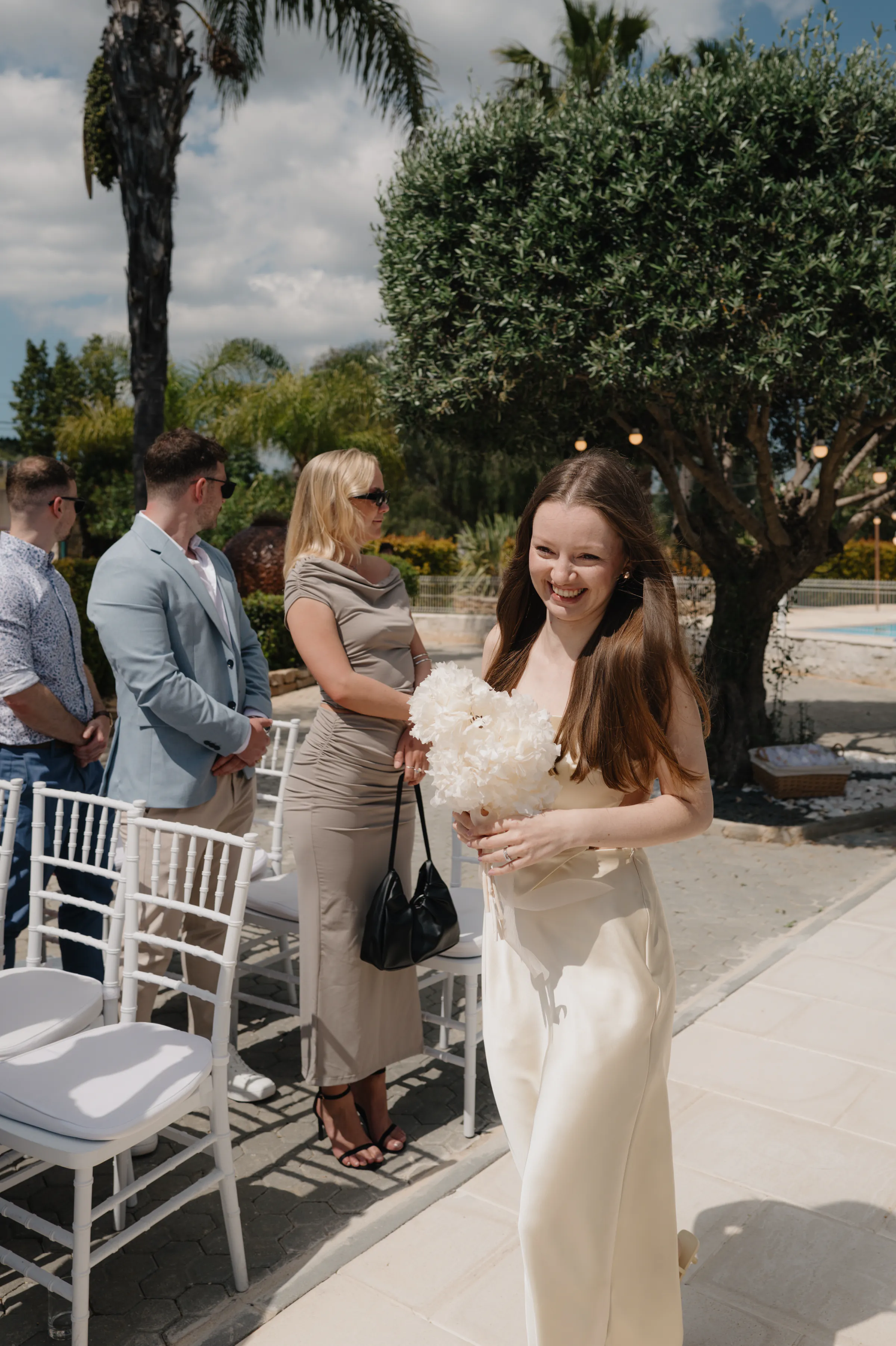 Woman in white dress holding a bouquet during an outdoor ceremony beside guests and white chairs.
