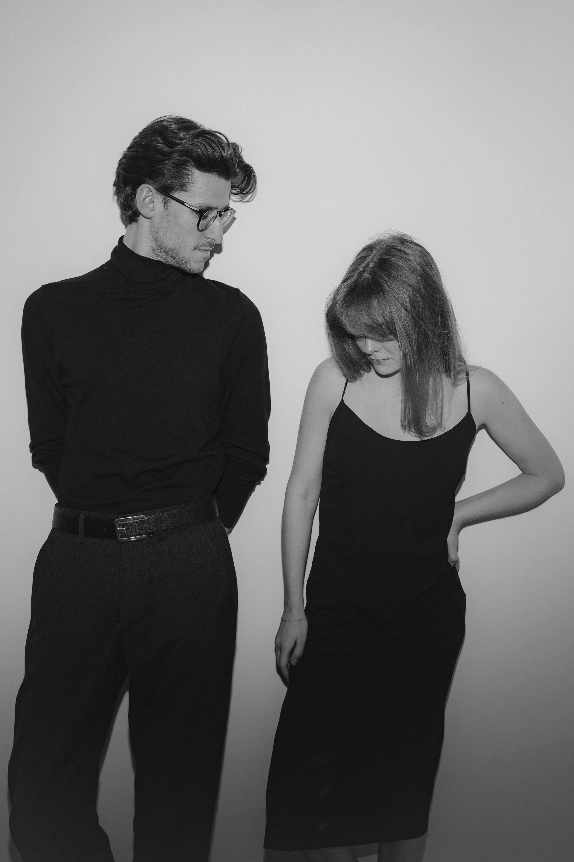 Black-and-white couple portrait in dark outfits against a plain studio backdrop, softly posed and looking downward.