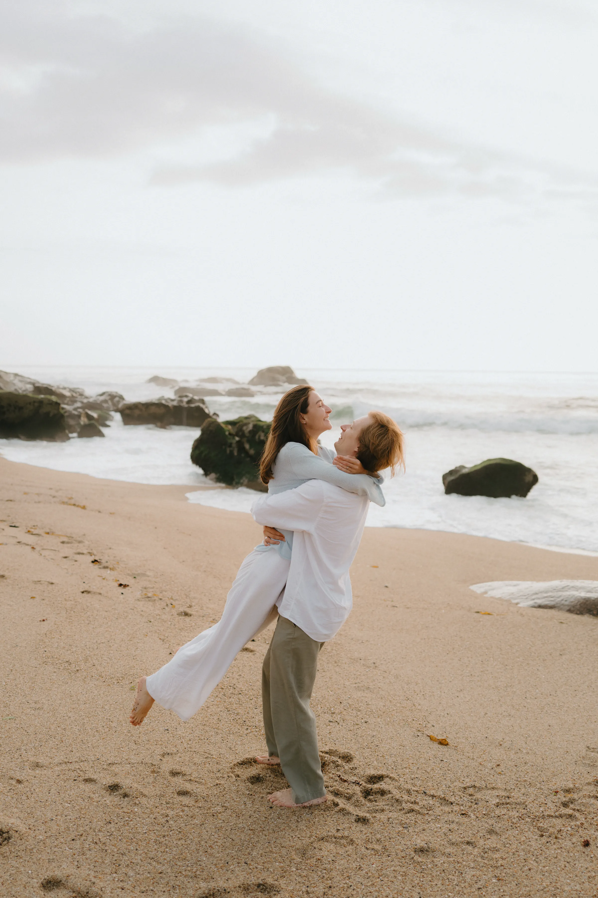Couple during a seaside session, one lifting the other on a sandy beach near rocks and soft surf.