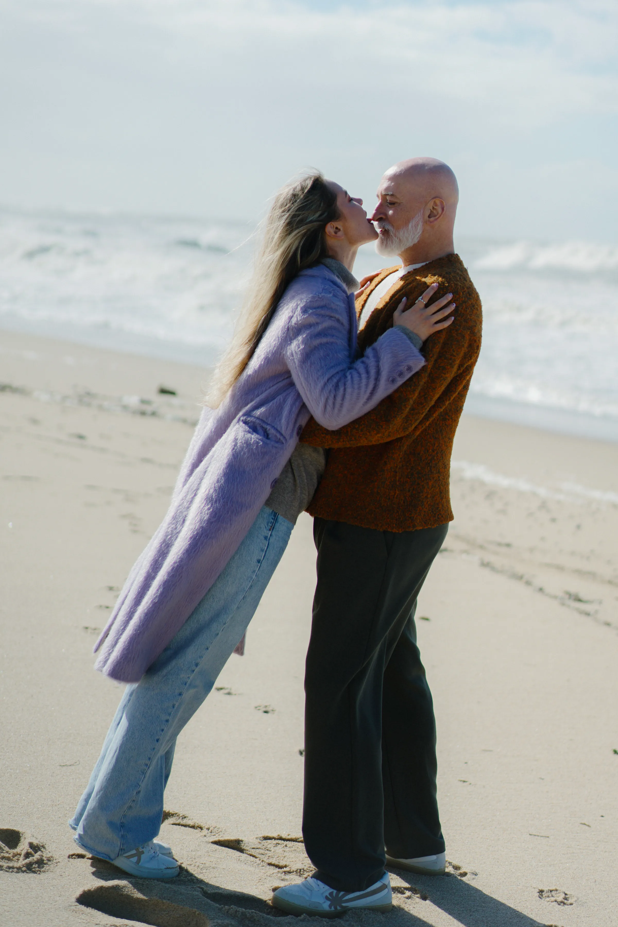 Couple kissing during a seaside outdoor session on a sandy beach with soft light and waves behind them.