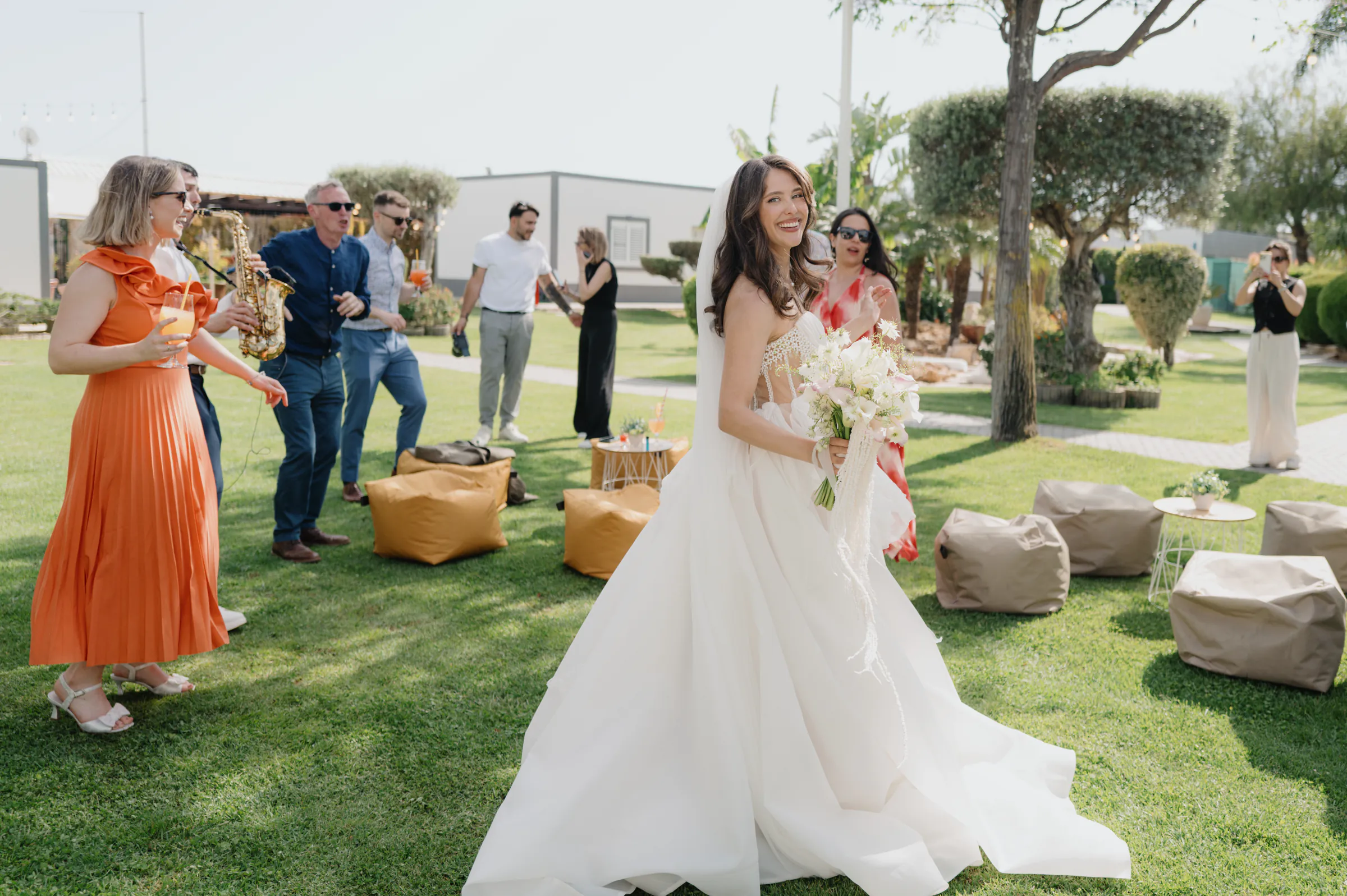 Bride with bouquet during an outdoor garden wedding session, with guests gathered on the lawn in soft light.