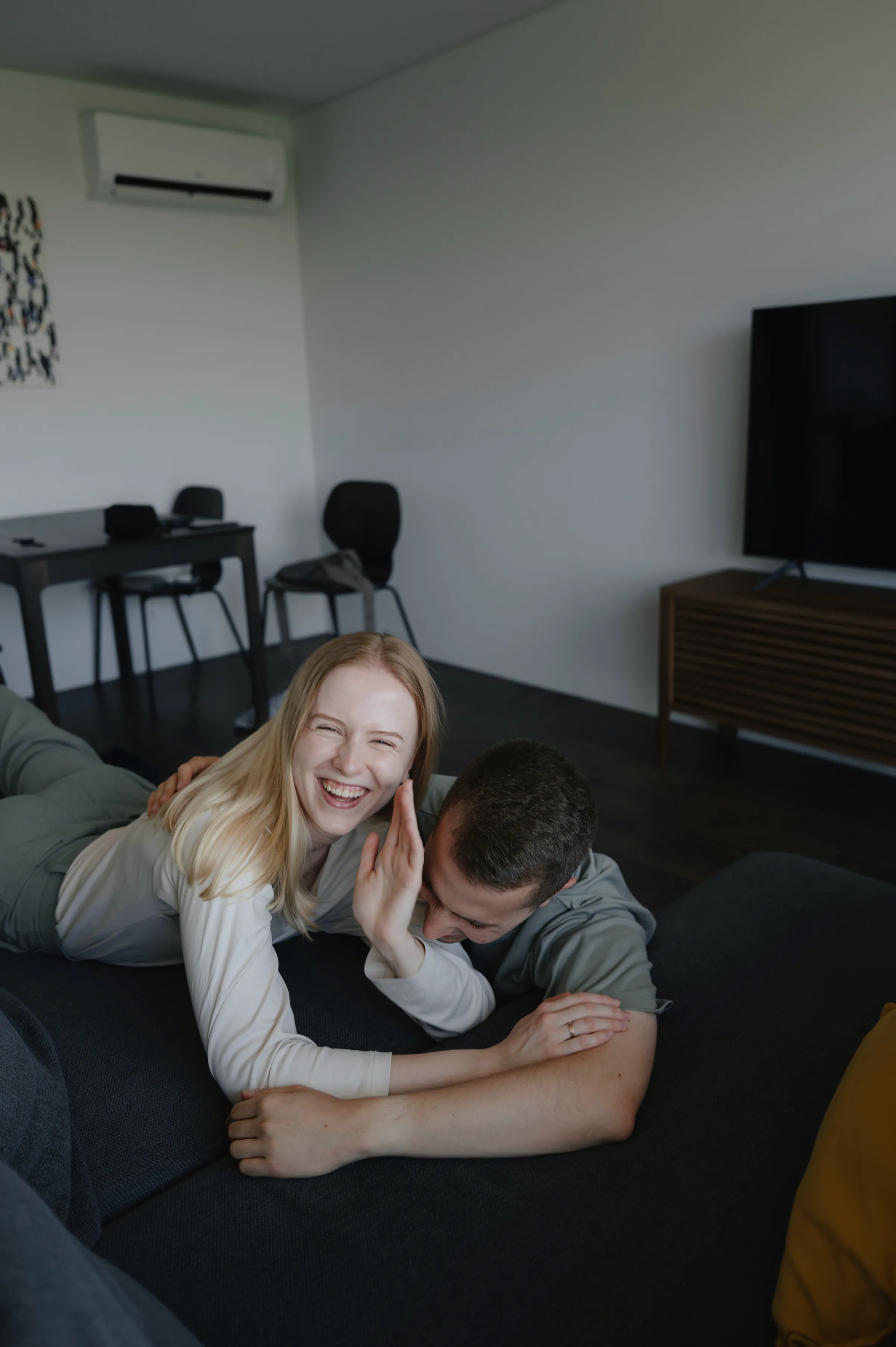 Couple lounging closely on a dark couch during a relaxed home session in soft indoor light.