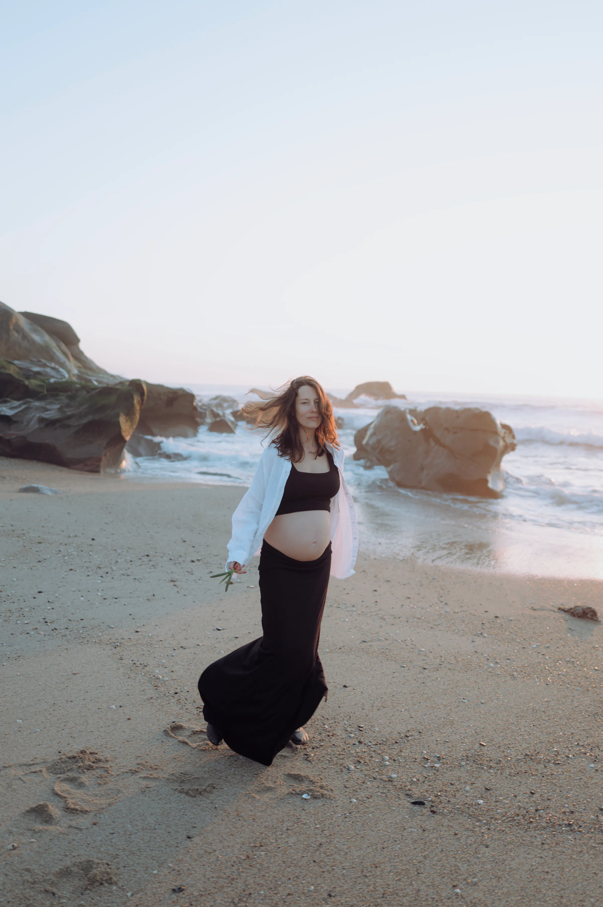 Seaside portrait of a person in a black dress and white shirt standing among rocks in soft light.